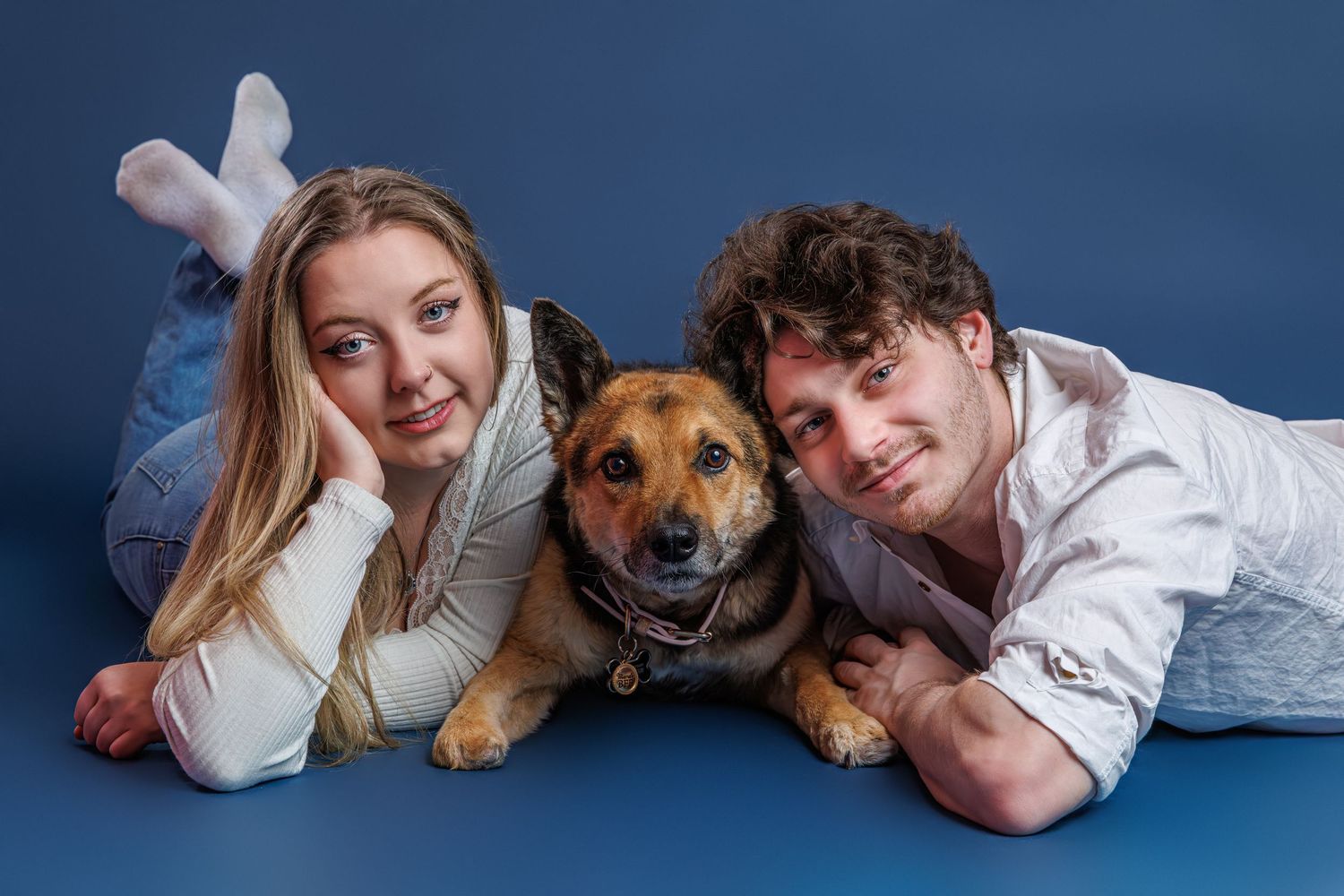 A smiling couple lies on a blue floor with a brown dog between them in a studio portrait photo.