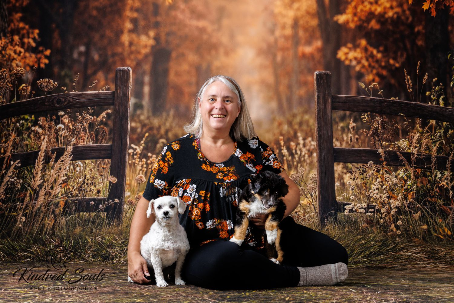 Woman sitting outdoors with a small white dog and a calico cat, surrounded by autumn foliage and a rustic wooden fence.