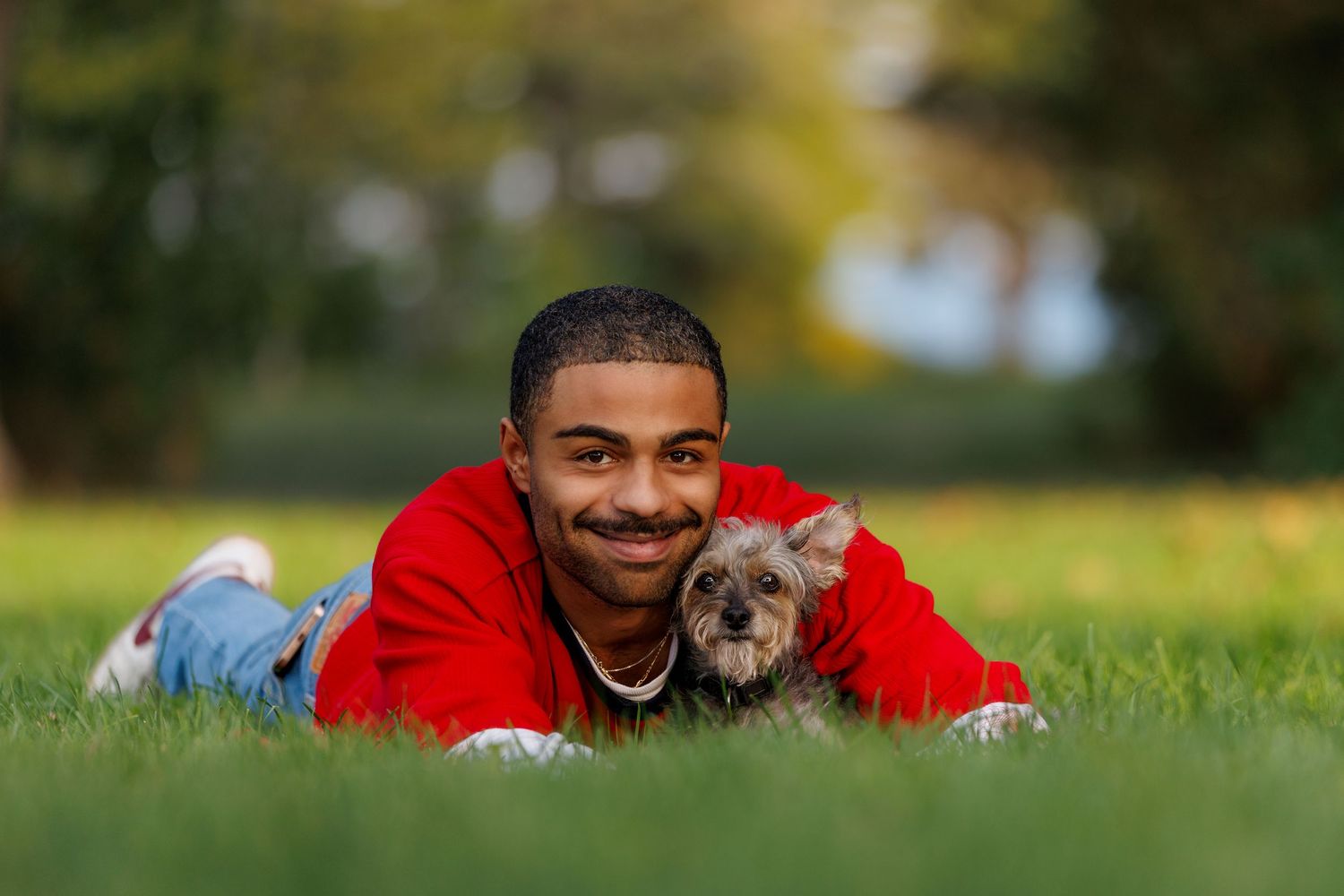 A smiling young man in a red hoodie lies on green grass with a small Yorkshire Terrier dog.
