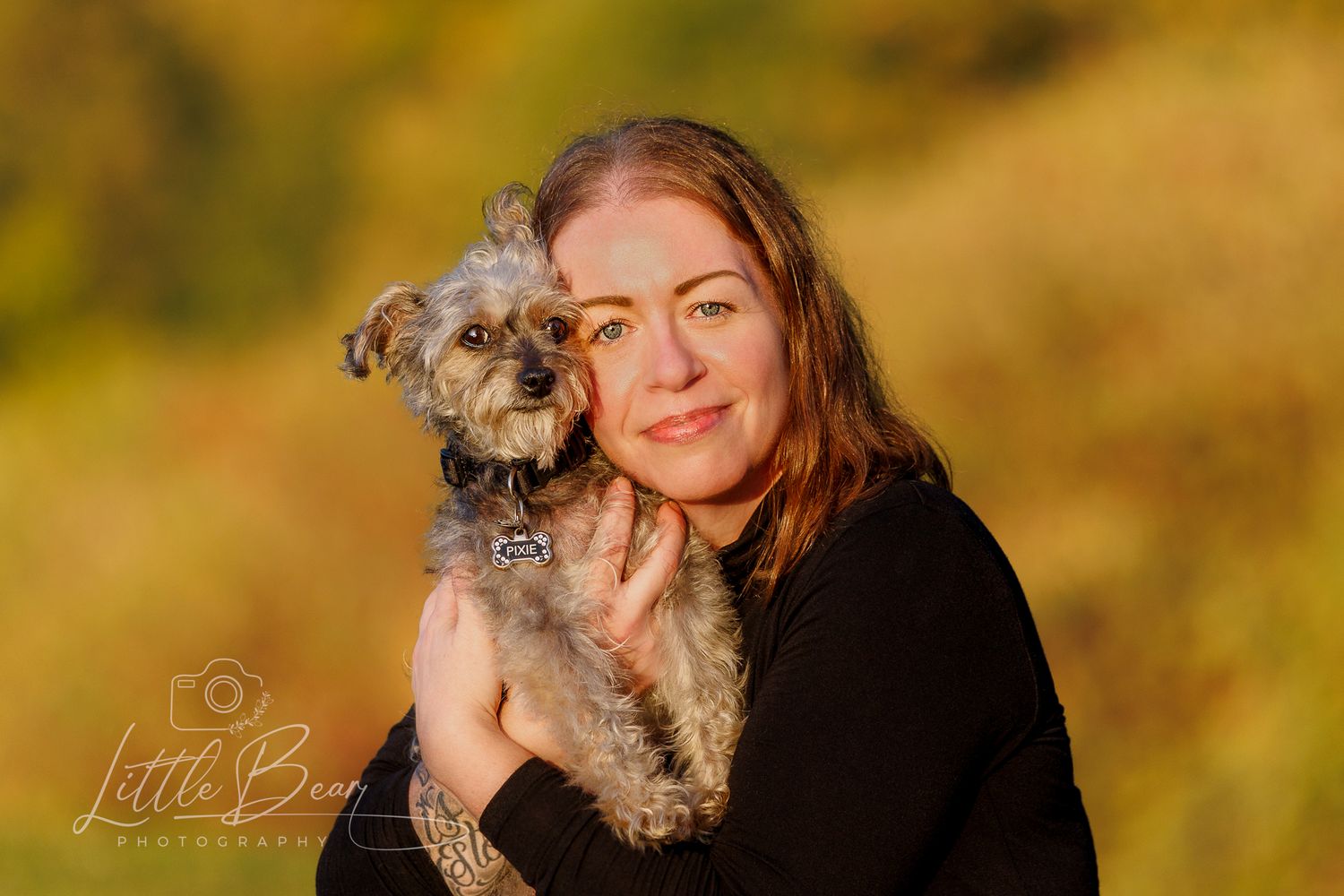 Woman holding a small gray schnauzer dog outdoors with golden autumn foliage in the background.