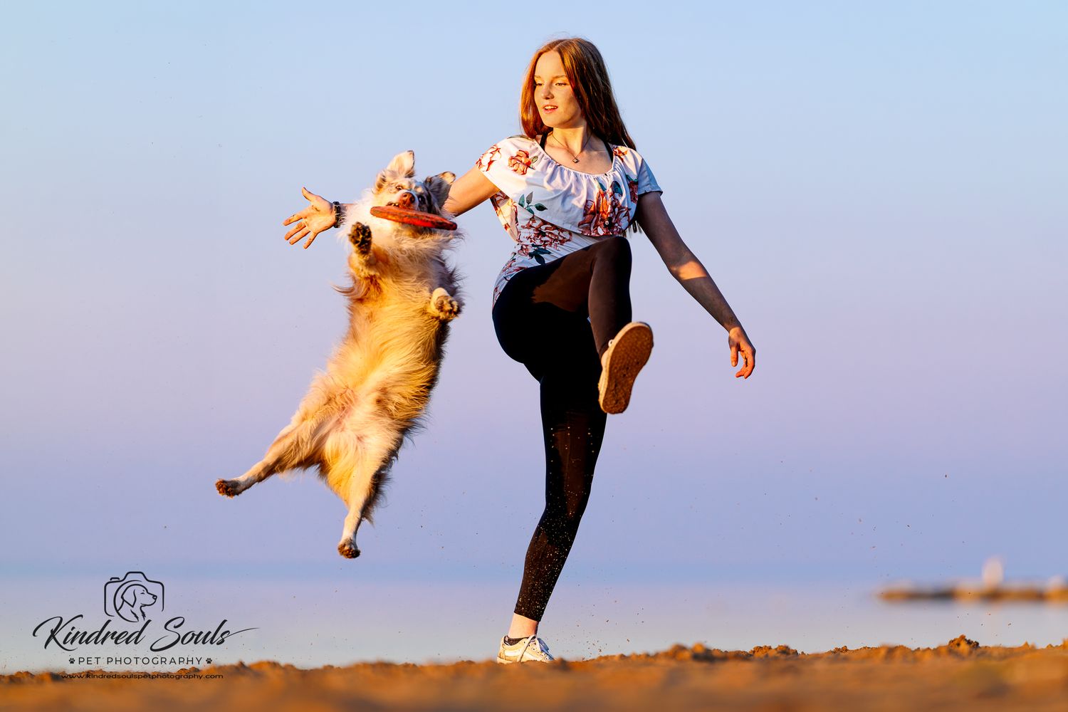 A woman in a white top and black leggings plays fetch with a jumping Collie dog on a sunny beach.