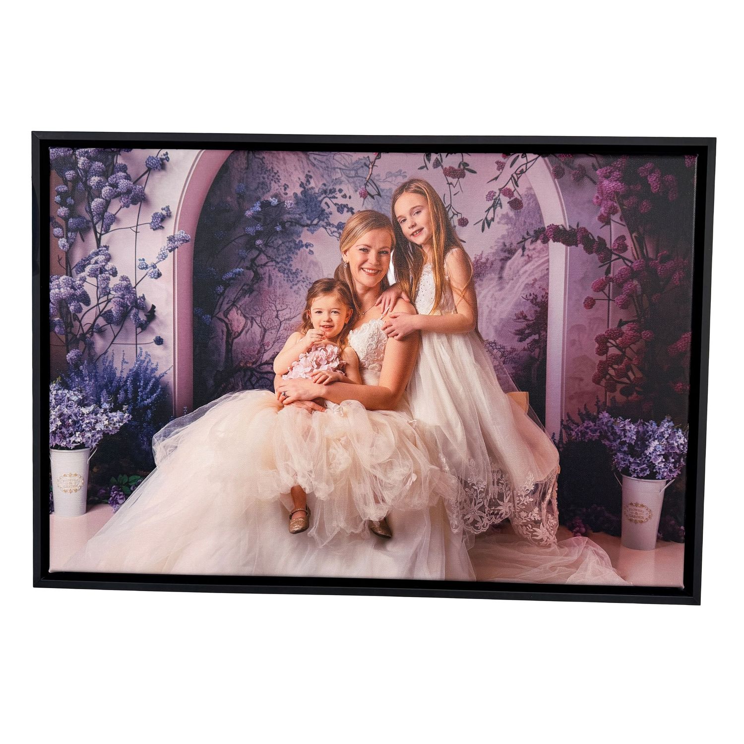 Framed portrait of a mother and two daughters in white dresses posed in a floral garden setting.