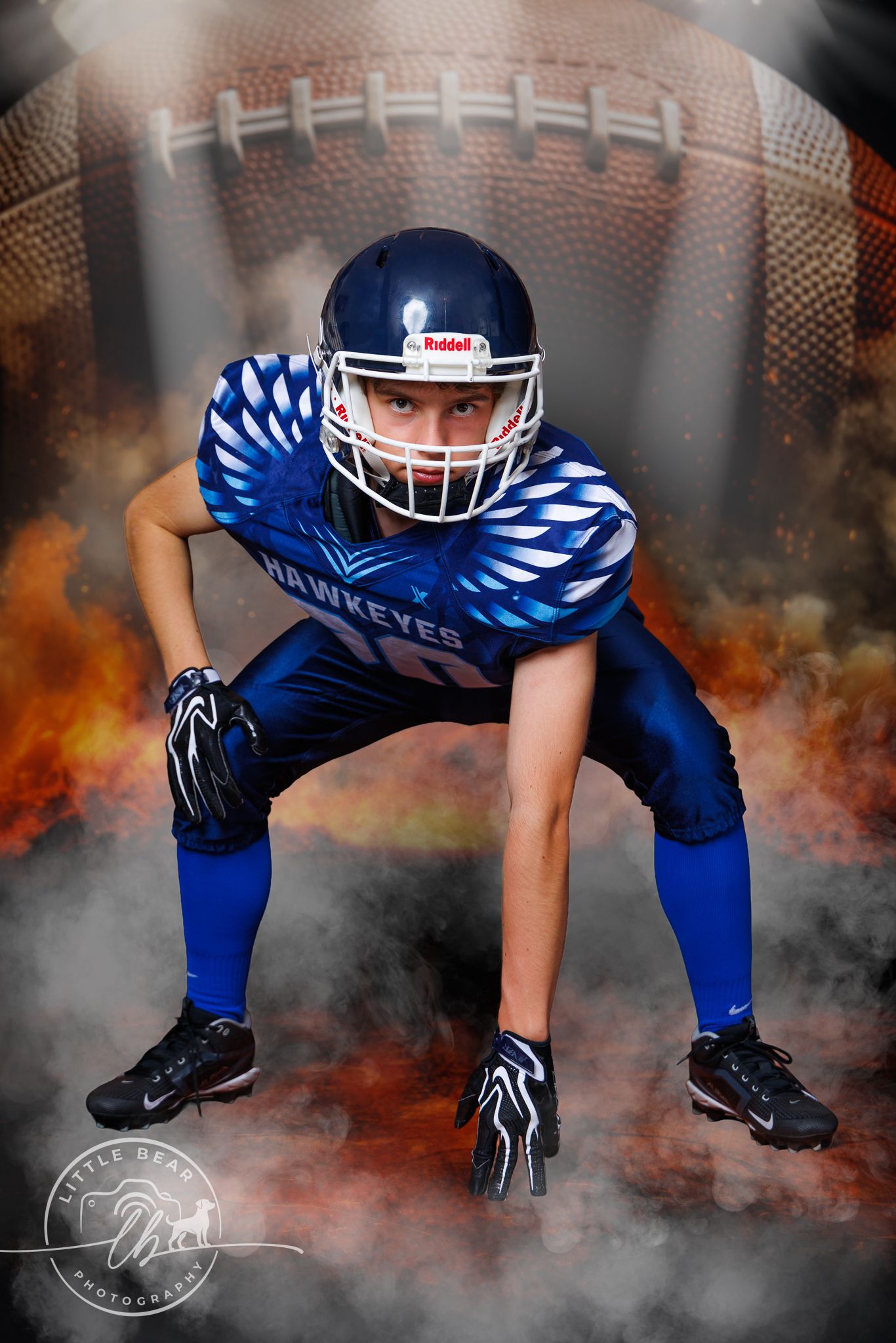 Young Hawkeyes football player in blue uniform crouches in ready stance with football and fire backdrop.