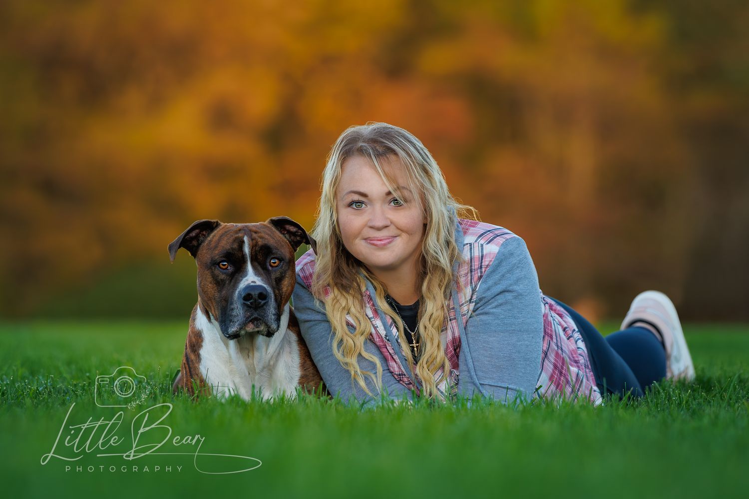 A blonde woman lies on green grass posing cheek-to-cheek with her brindle Boxer dog in autumn light.