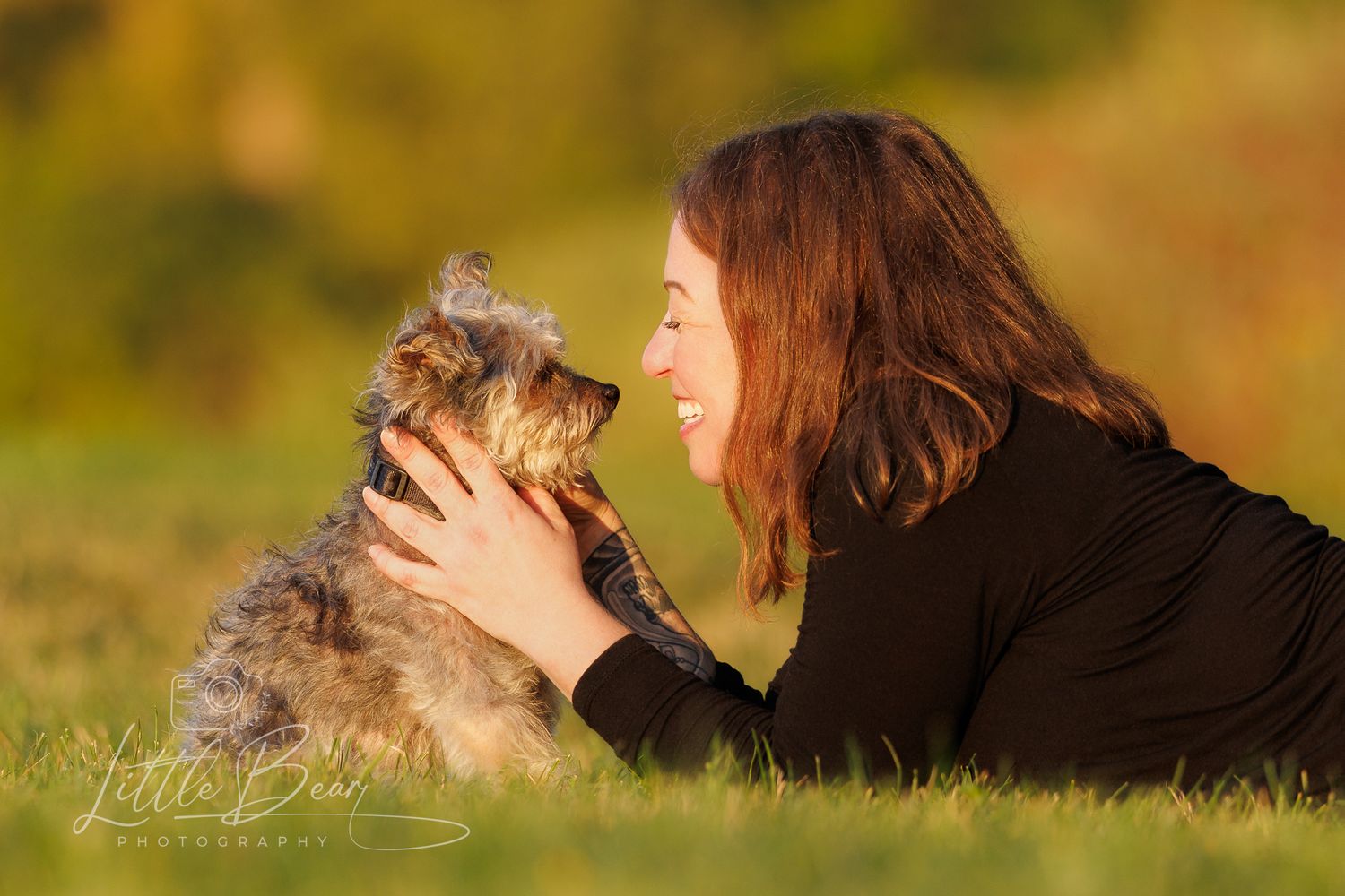 Woman in black top lying on grass face-to-face with a small fluffy dog outdoors in warm golden light.