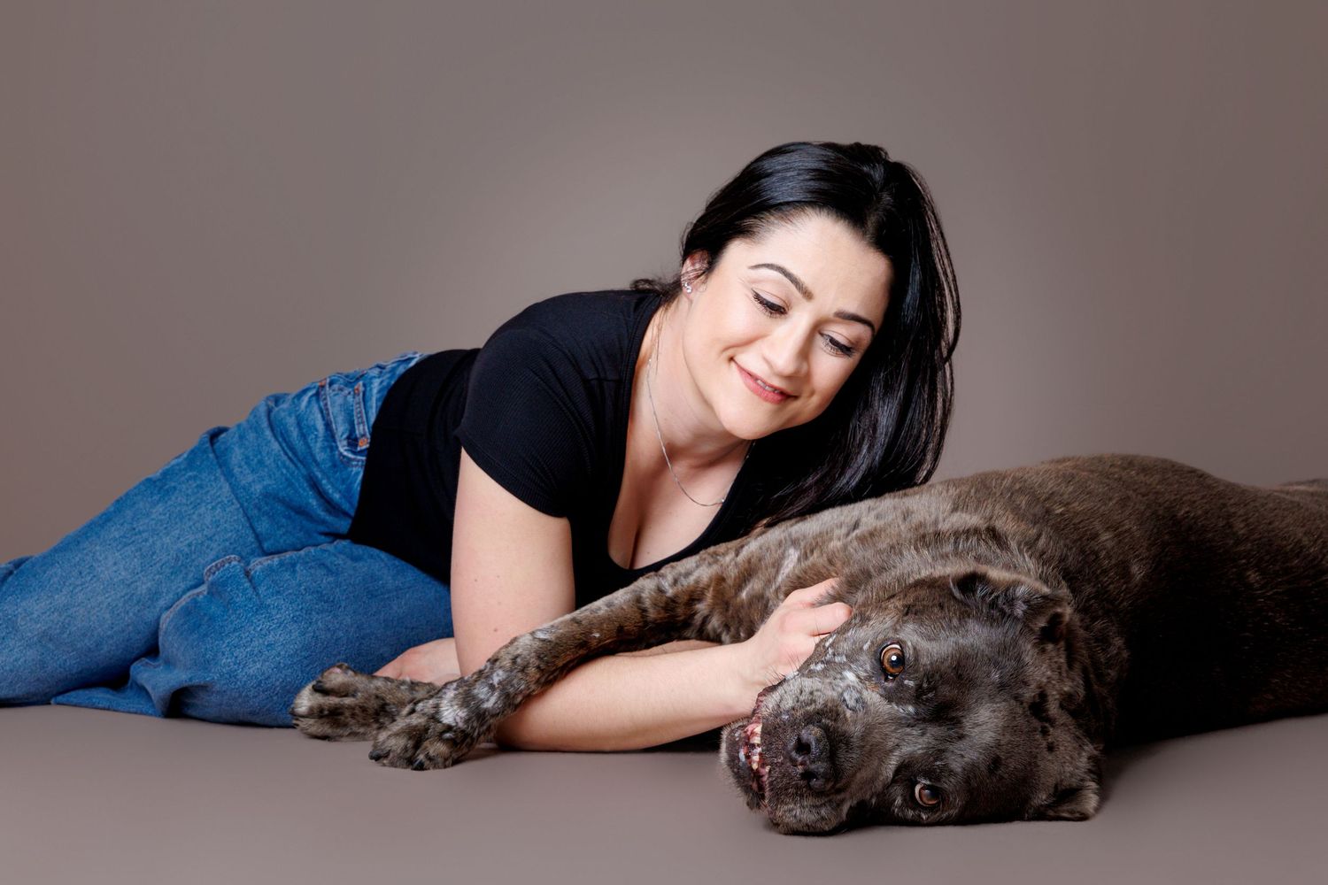 Woman in black shirt and jeans lying on floor smiling while embracing a gray brindle Cane Corso dog.