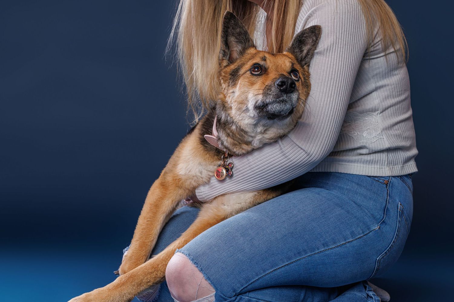 Woman in jeans holding a brown mixed-breed dog on her lap against a dark blue background.