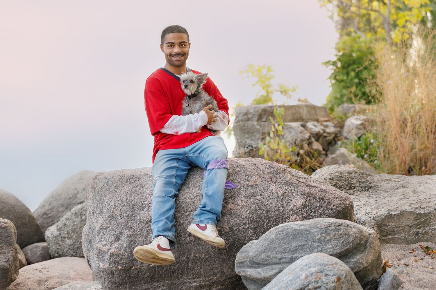A smiling man in a red sweater sits on rocks holding a small gray Yorkshire Terrier dog.