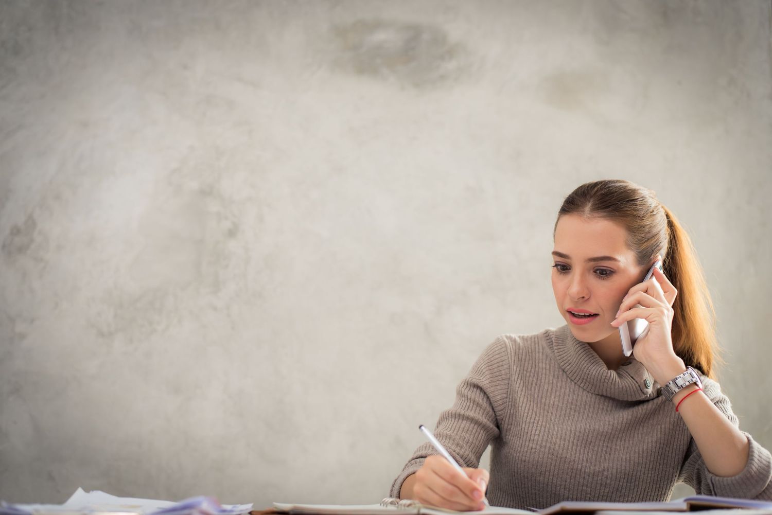 A young woman talks on the phone while taking notes at a desk against a gray wall.