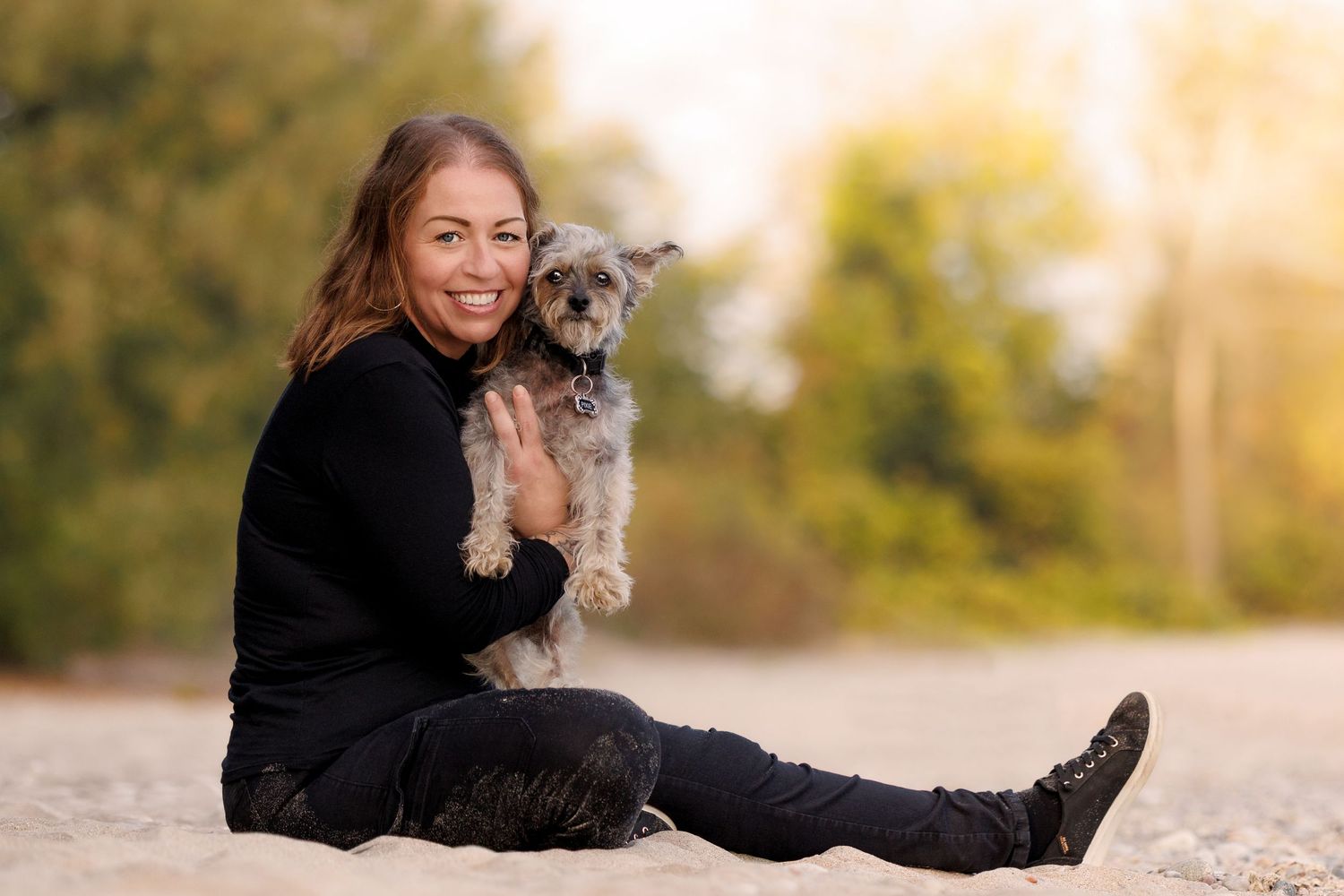 A smiling woman in black sits on sand holding a small gray Yorkshire Terrier dog outdoors.