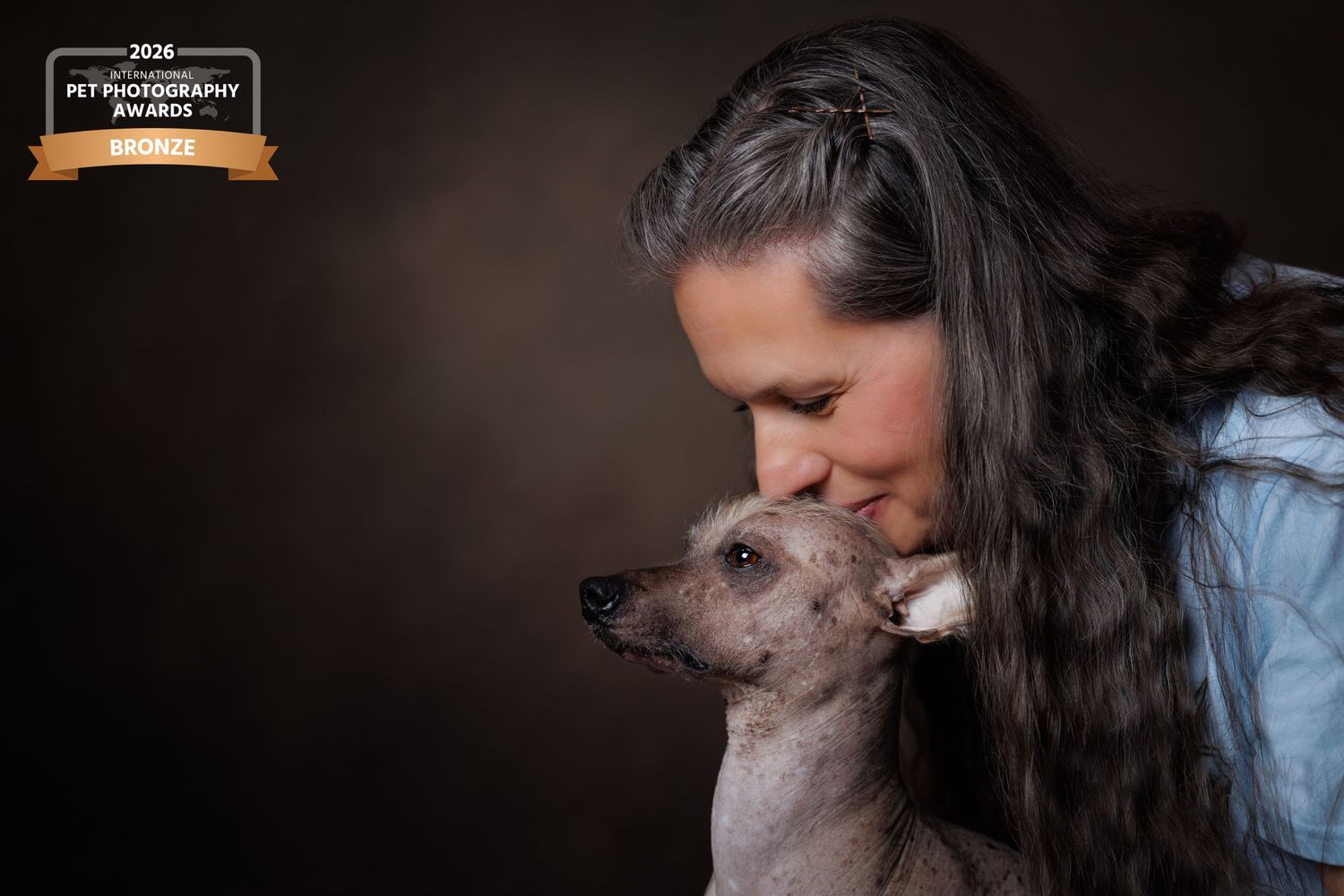 Woman with long dark hair kissing a Chinese Crested dog against a dark background, award-winning pet photography.