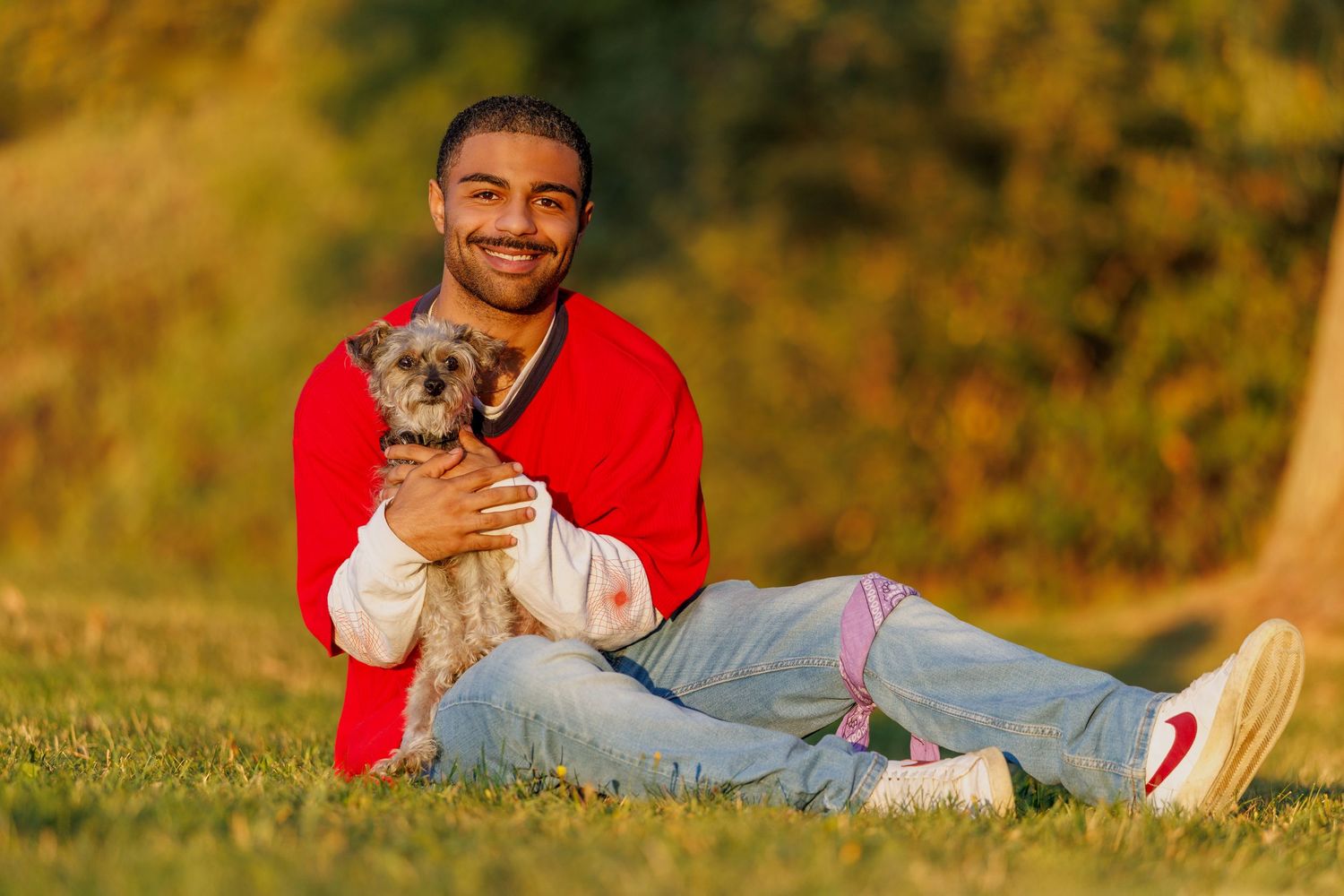 Smiling young man in red jacket sitting on grass holding small terrier dog outdoors.