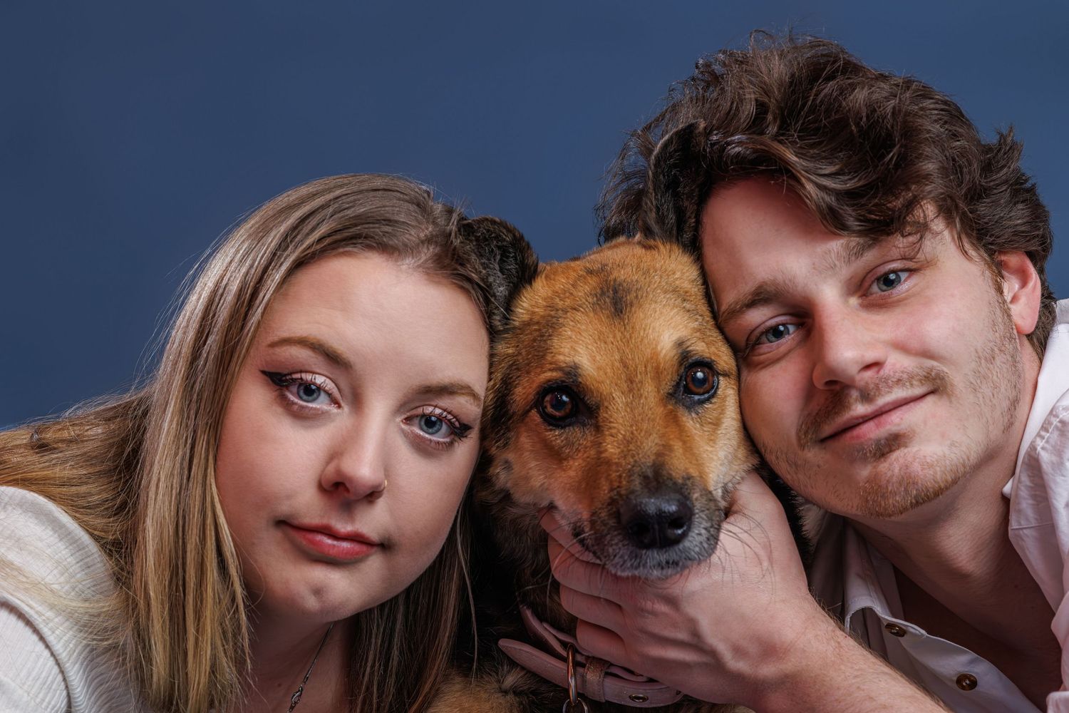A young couple posing with their brown mixed-breed dog against a blue studio background.