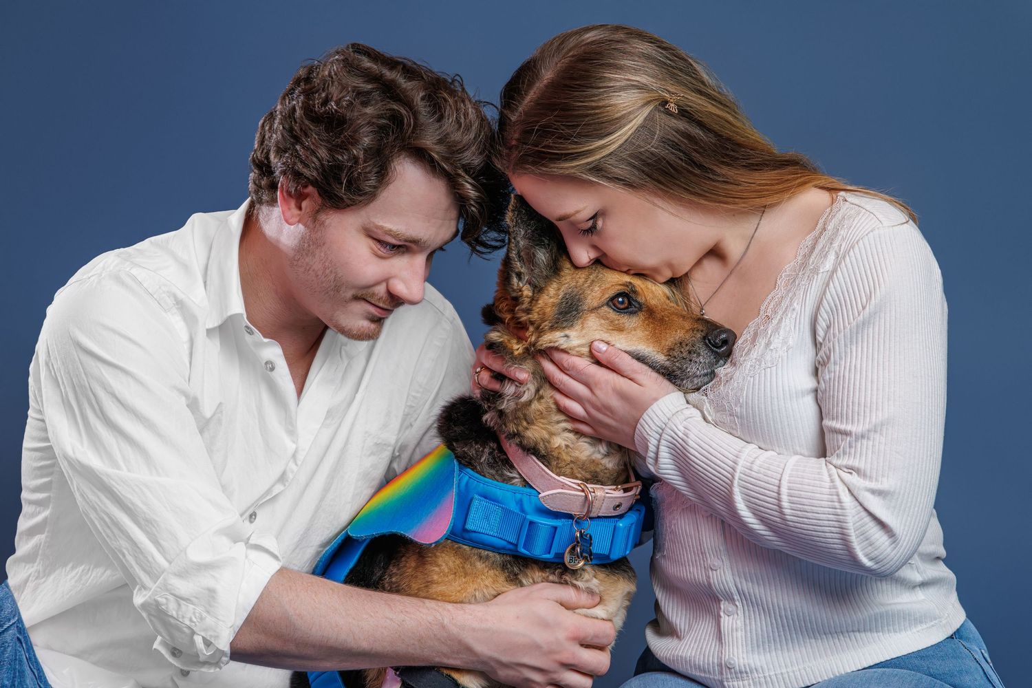 A couple leans in to kiss their small dog dressed in a rainbow outfit against a blue background.