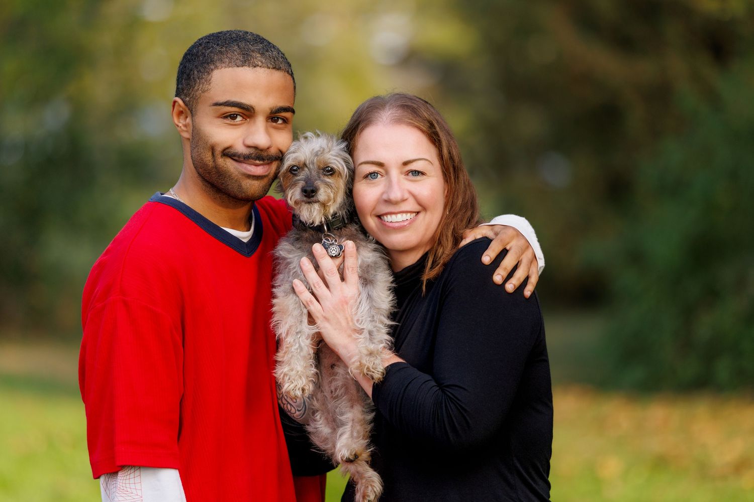 A smiling couple in a park holds a small gray terrier dog between them outdoors.