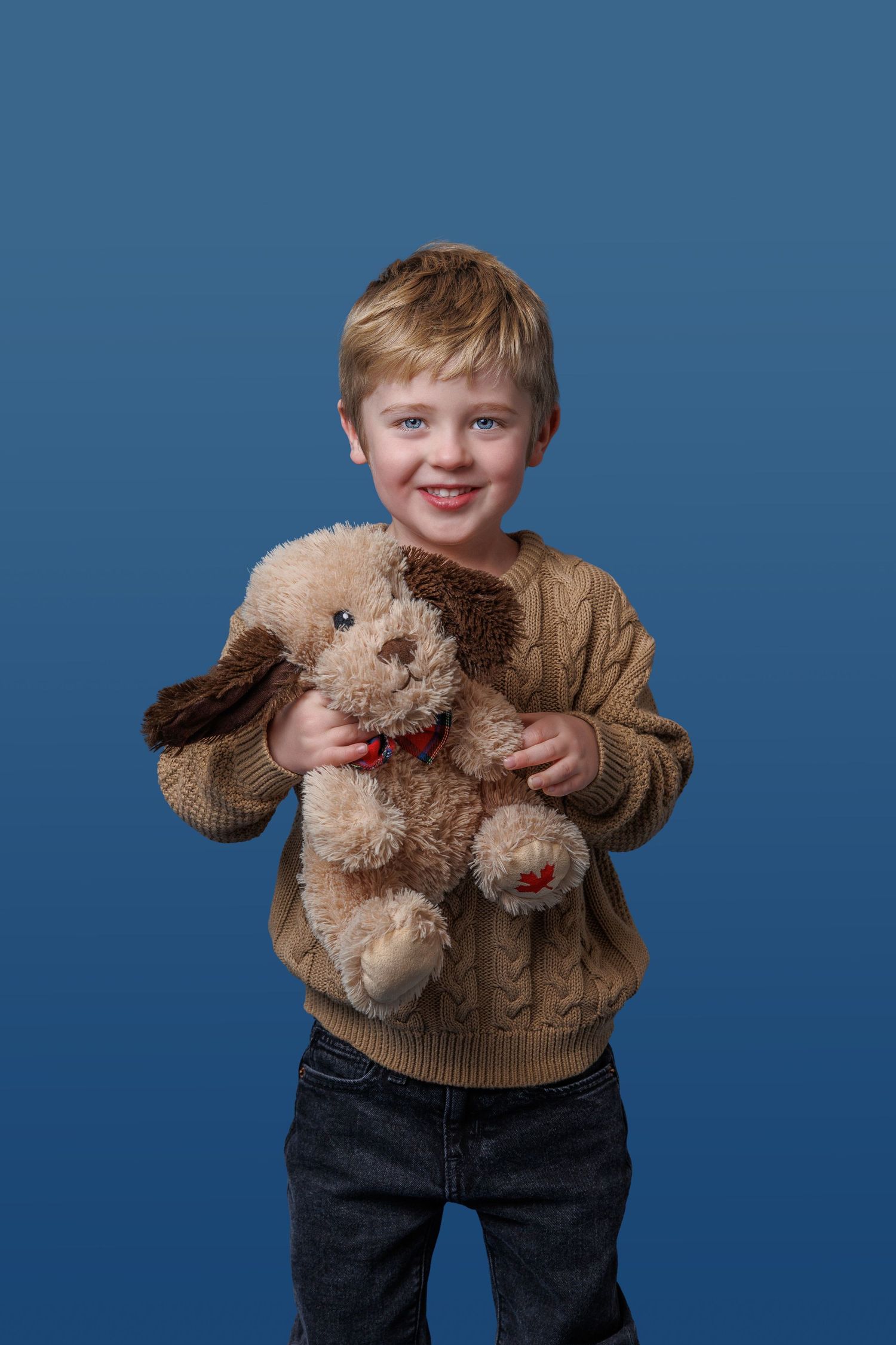Smiling young boy holding a stuffed toy dog on a blue studio background.