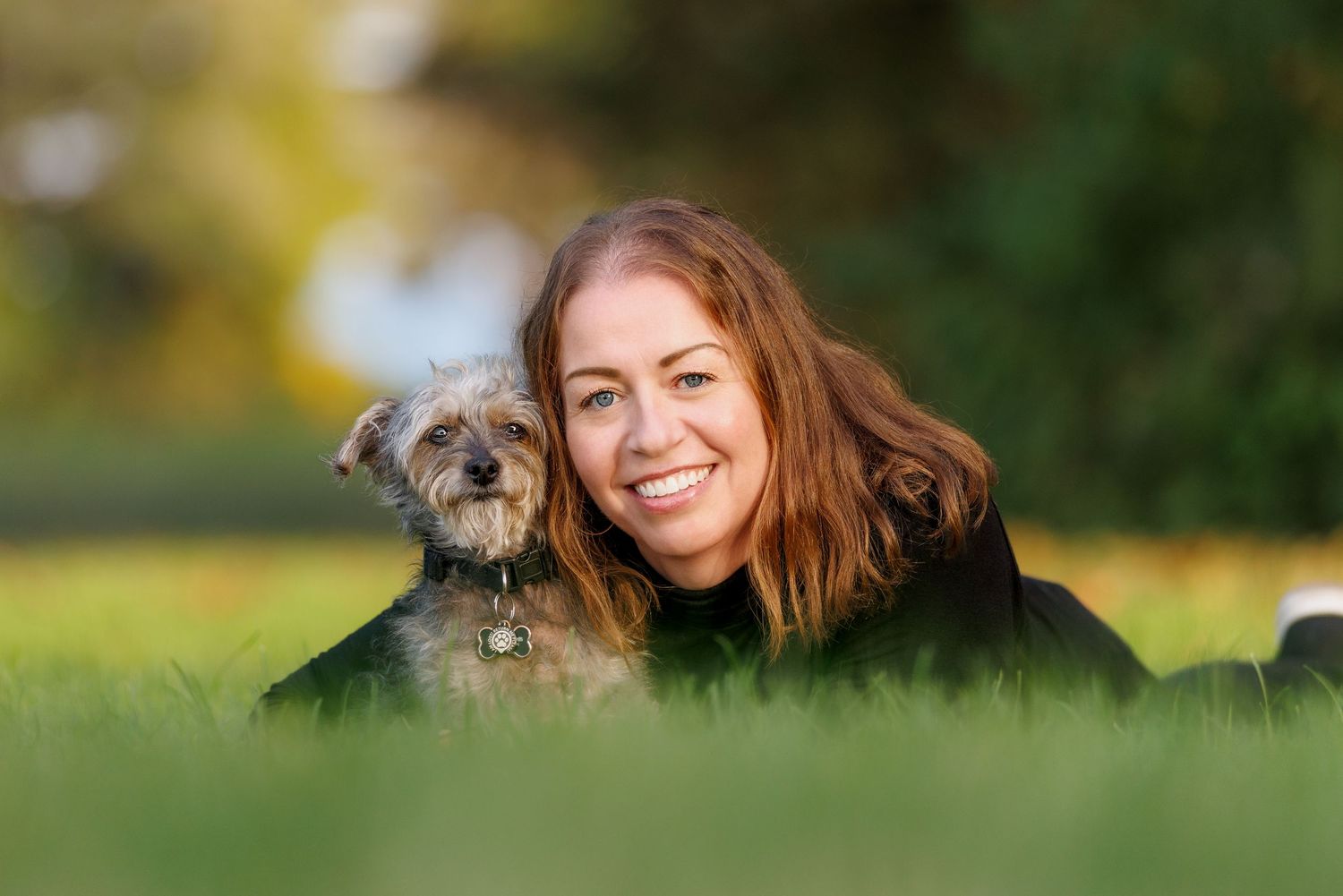 A smiling woman with red hair lies on green grass with her small gray terrier dog.