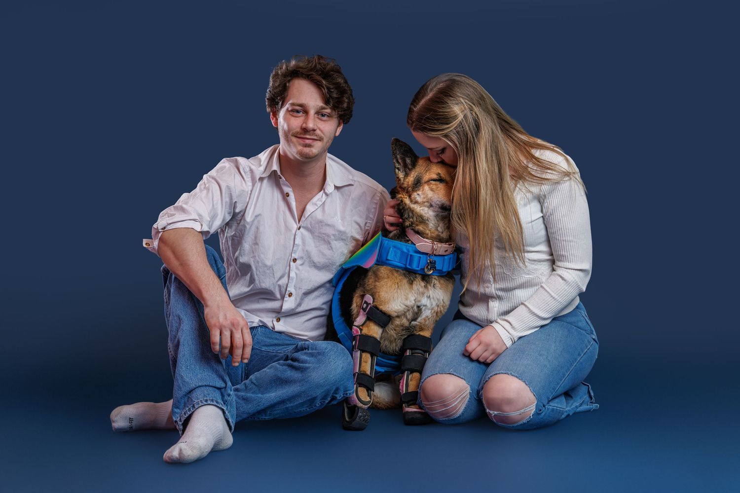 A couple sitting on the floor with a German Shepherd dog wearing a blue harness against a dark blue background.