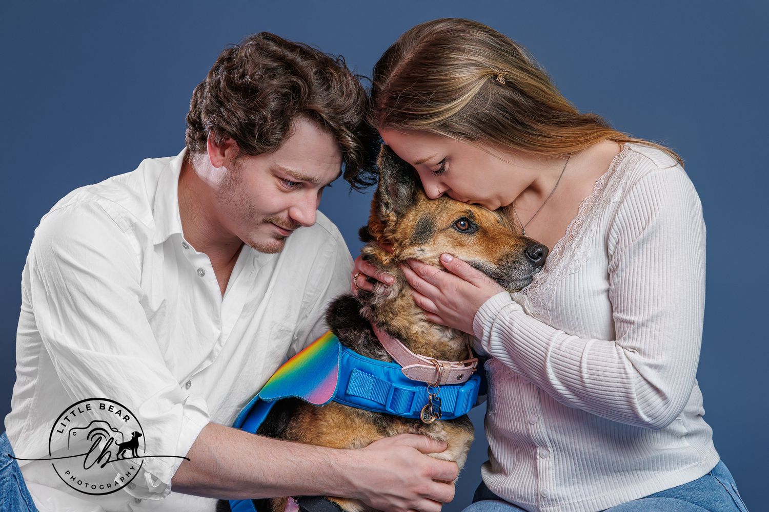 A couple lovingly nuzzles their German Shepherd dog wearing a rainbow outfit during a professional studio photo session.