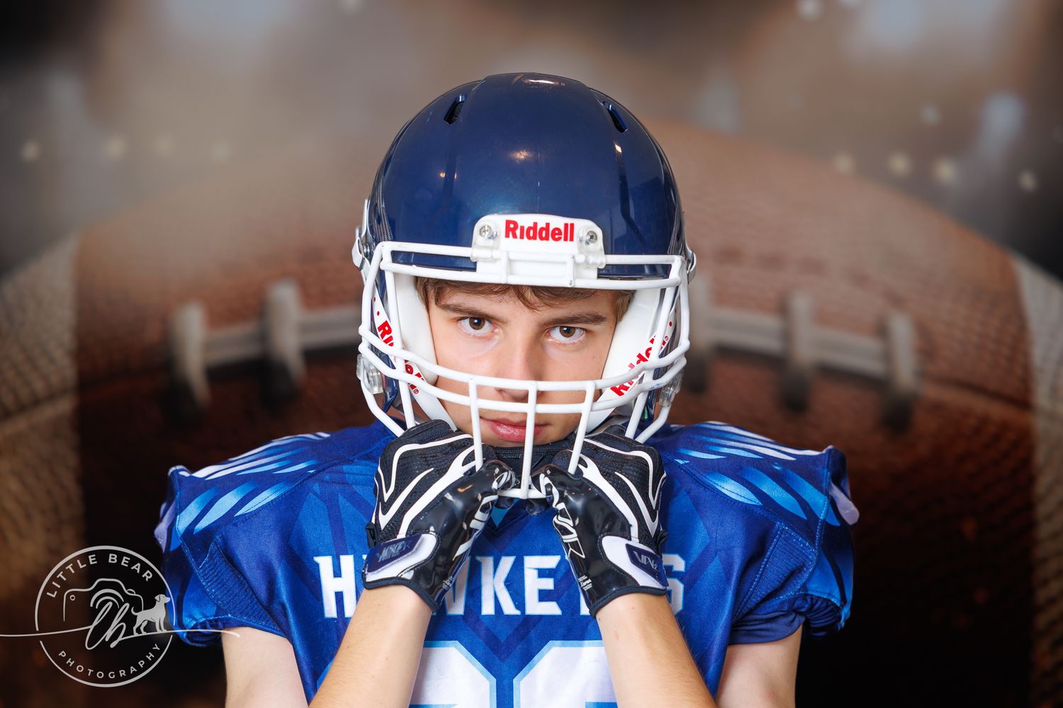 Young football player in blue Riddell helmet and jersey poses seriously with football background.