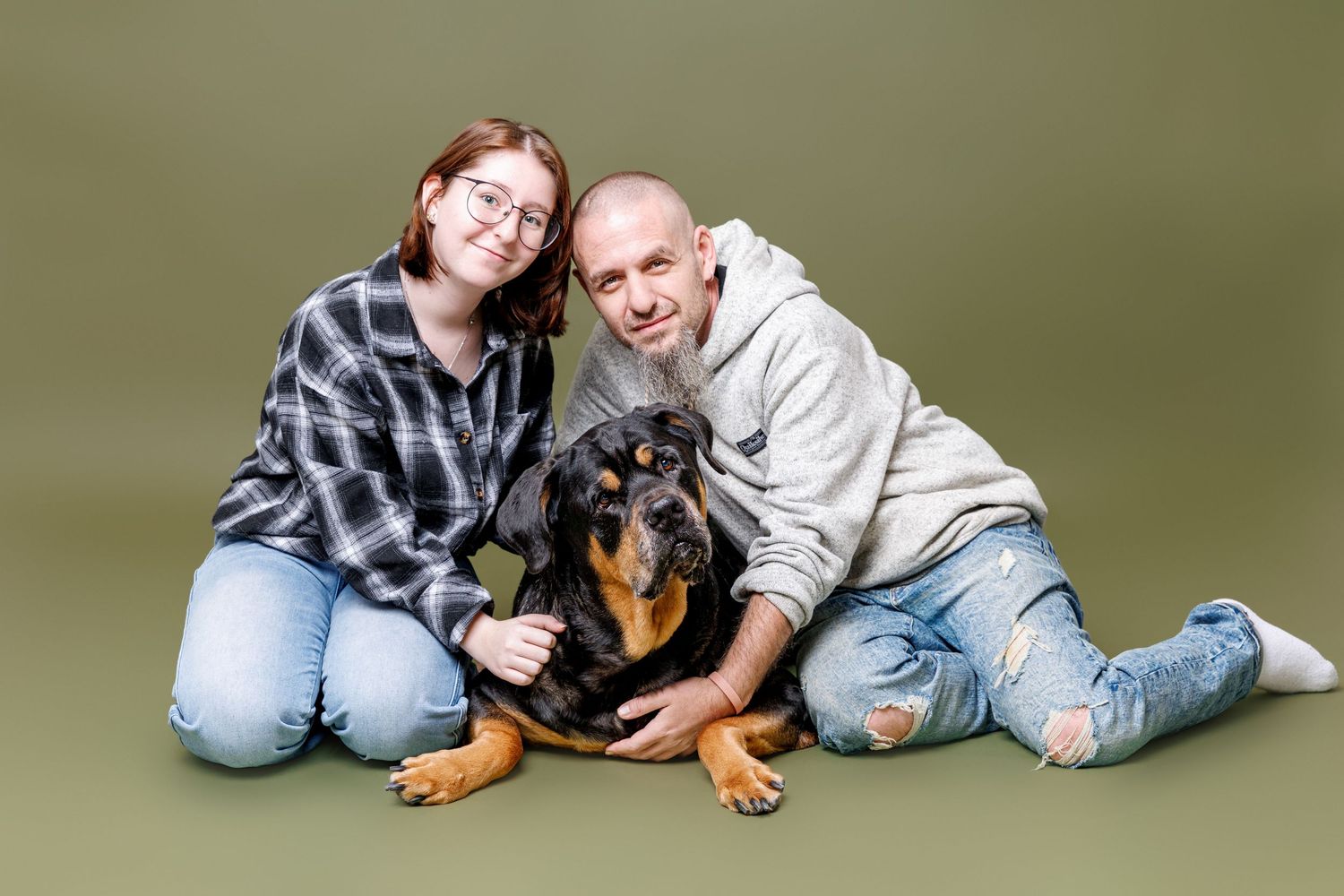 Smiling couple sitting on green background with their Rottweiler dog between them.