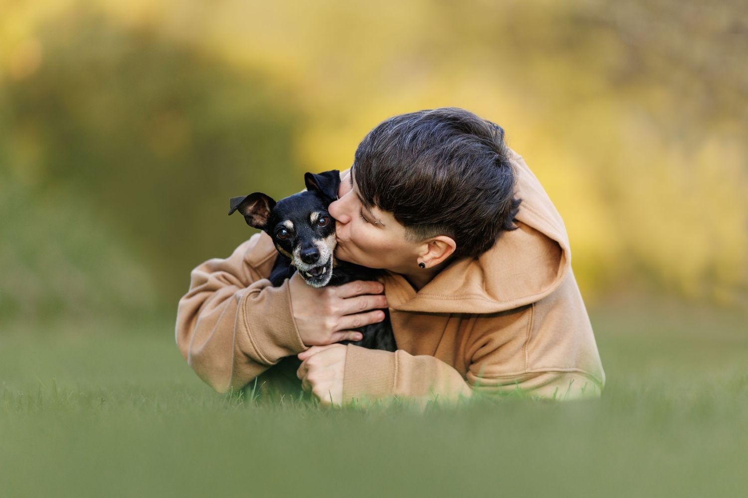 A young girl lying on grass lovingly kissing her small black dog.