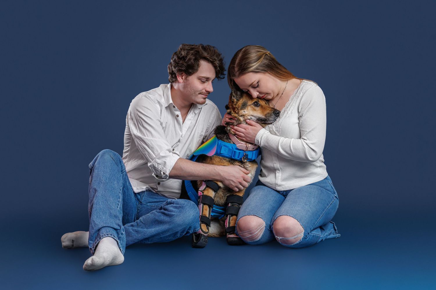Couple sitting on blue studio floor holding a small dog dressed in a blue outfit, smiling and bonding together.