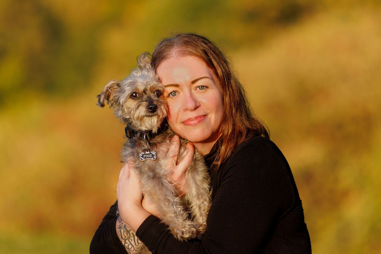 Smiling woman holding small gray terrier dog with autumn foliage in background.