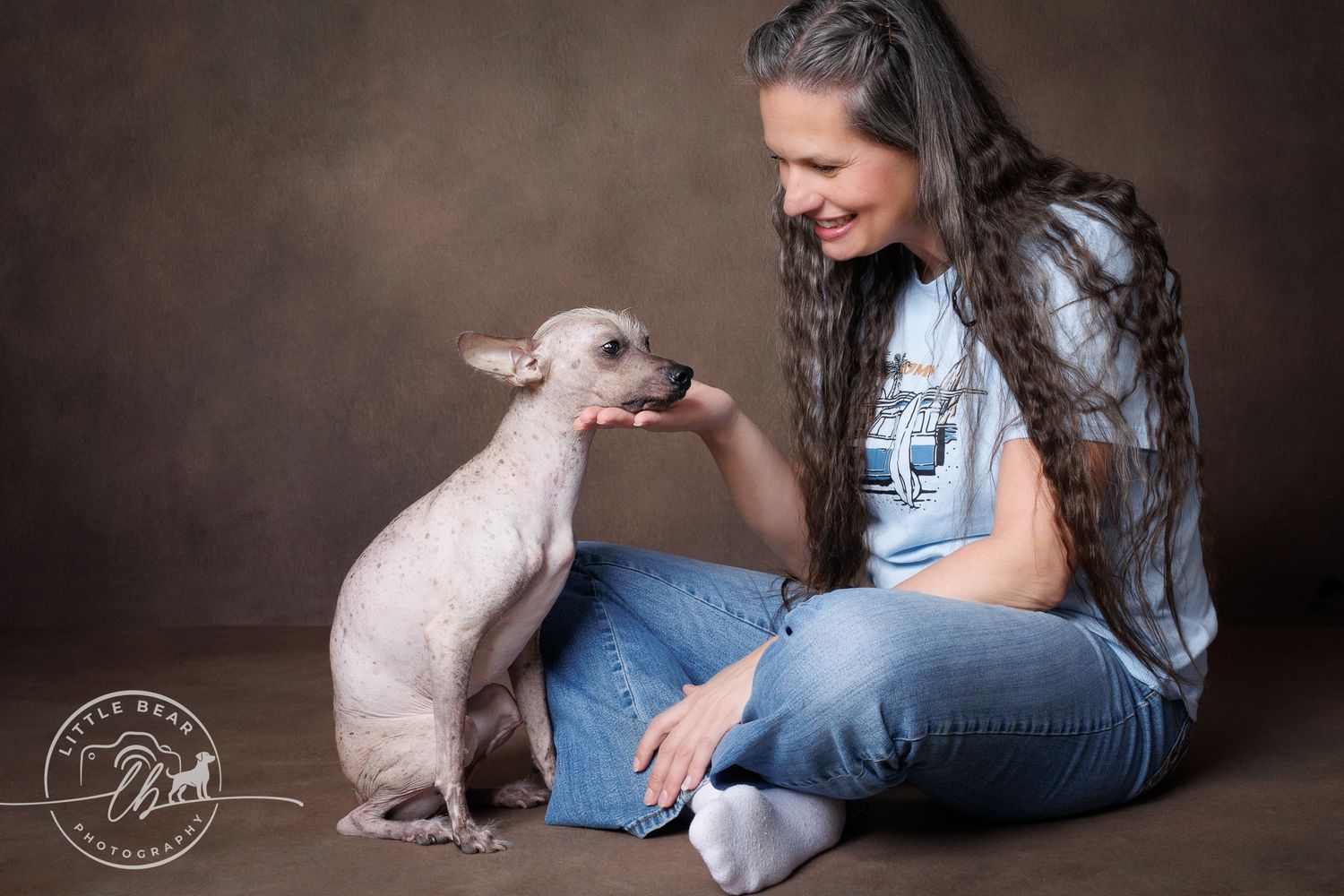 A woman with long hair smiles at a hairless dog licking her hand during a professional studio photo session.