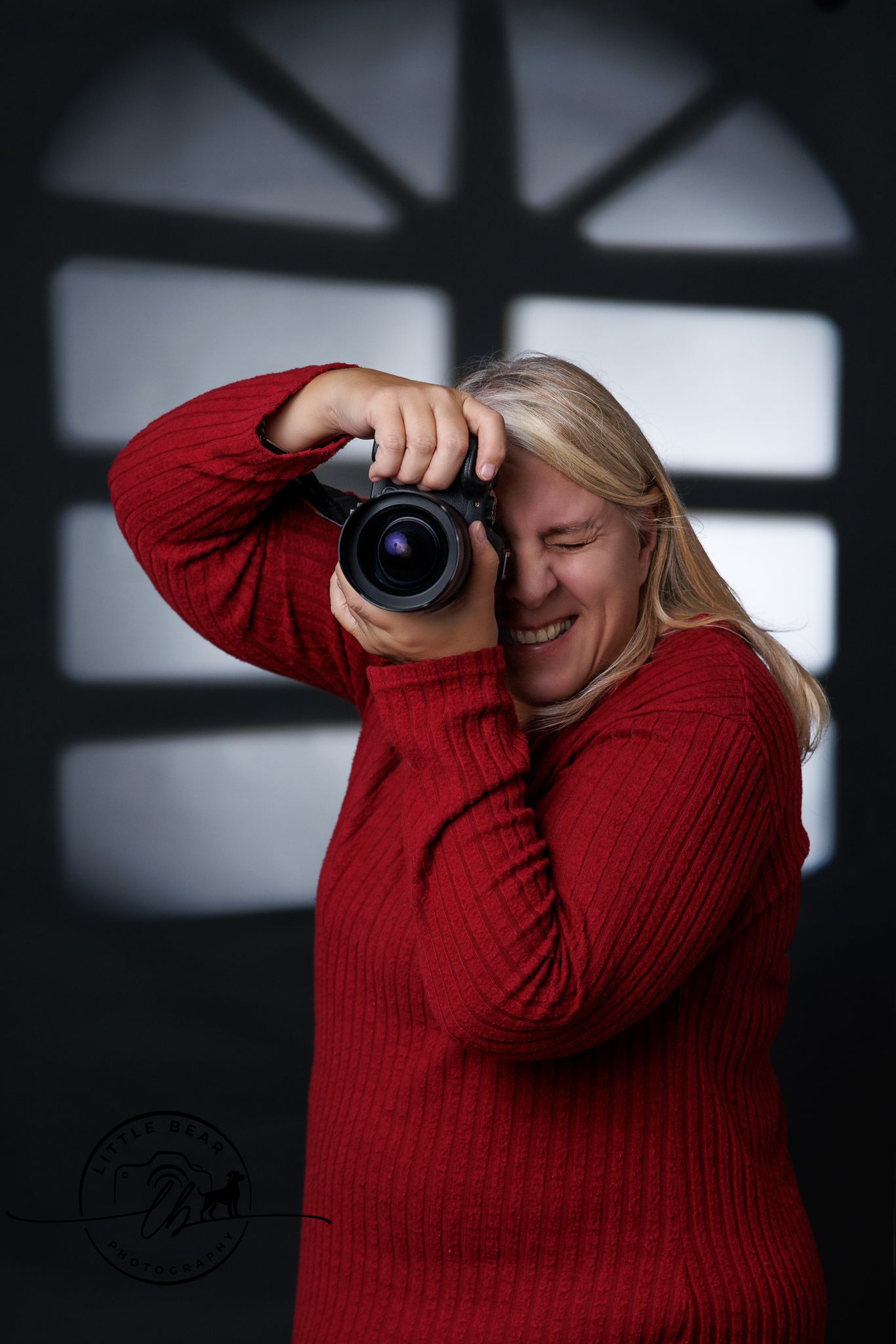 Blonde woman in red sweater laughing while looking through Canon R5ii camera viewfinder in studio.