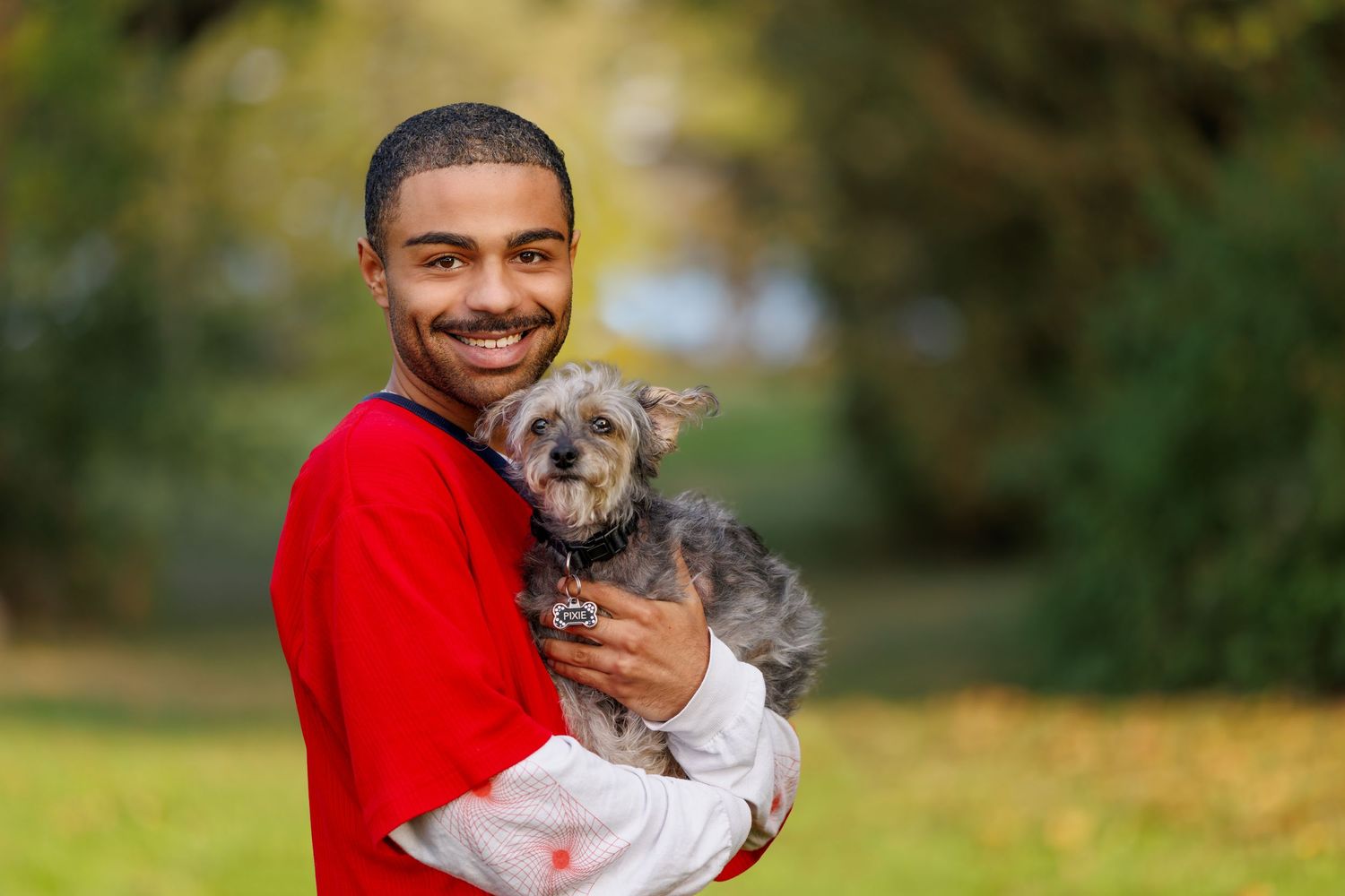 A smiling young man in a red sweatshirt holds a small gray terrier dog outdoors.