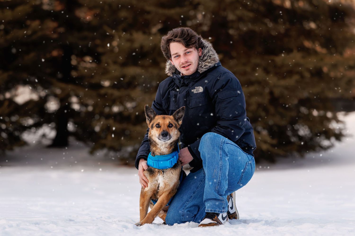 Person kneeling in snow with a dog wearing a blue bandana, surrounded by snowy trees in winter.