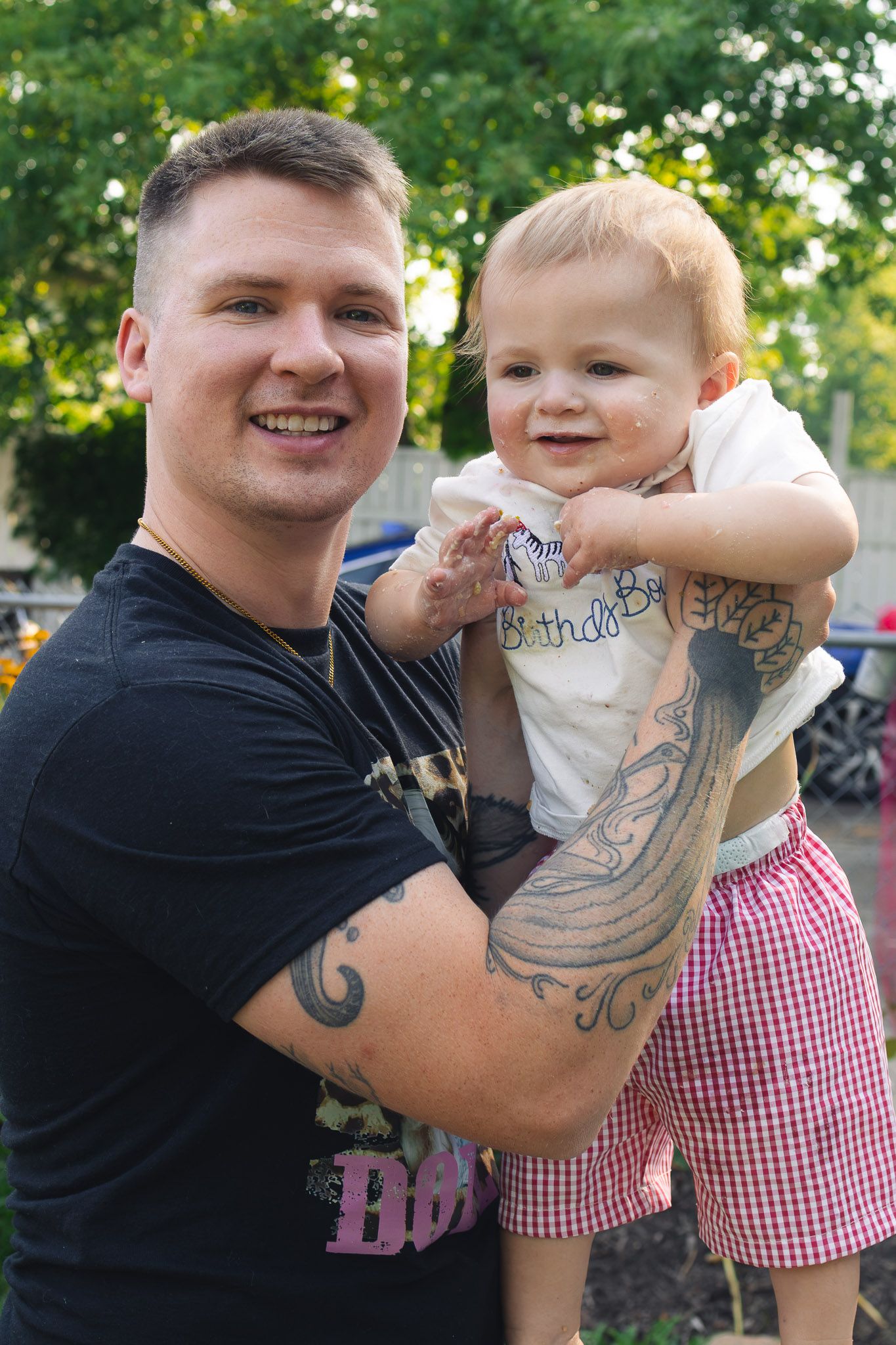 Smiling tattooed man in a black t-shirt holds a toddler in a birthday shirt and pink gingham shorts outdoors near green trees.
