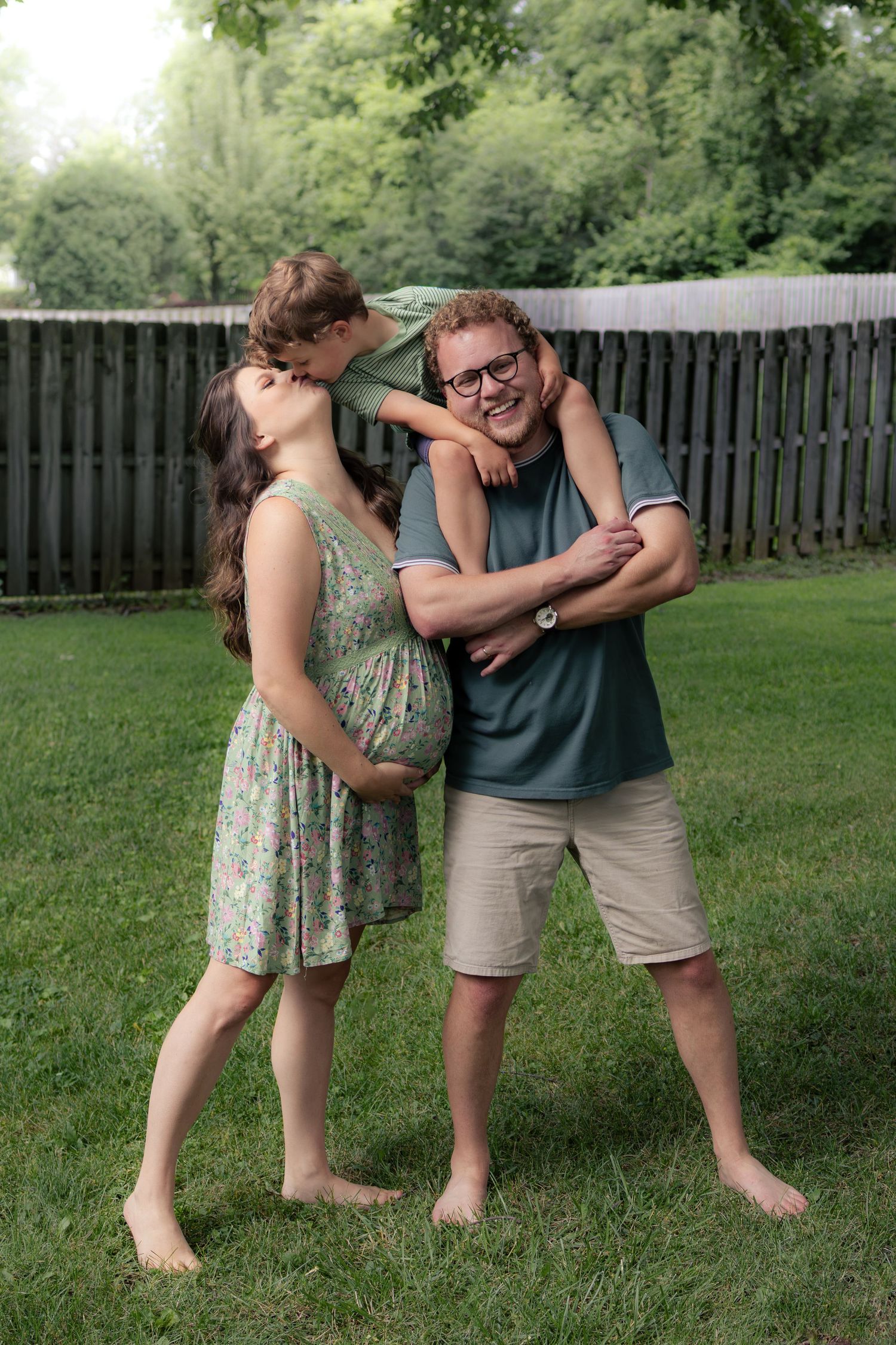 A pregnant woman and her partner share a laugh as their child climbs on dad's back outdoors.
