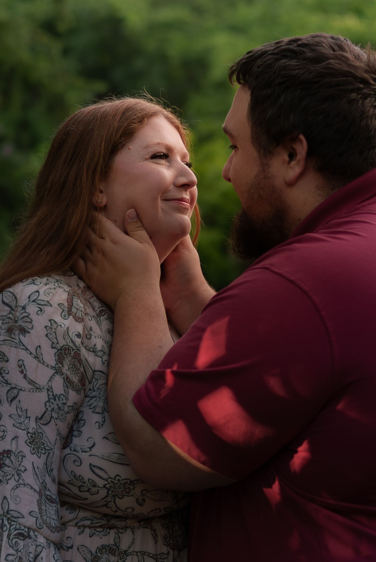 A couple shares a tender moment outdoors, man gently holding womans face as she smiles up at him among green foliage.