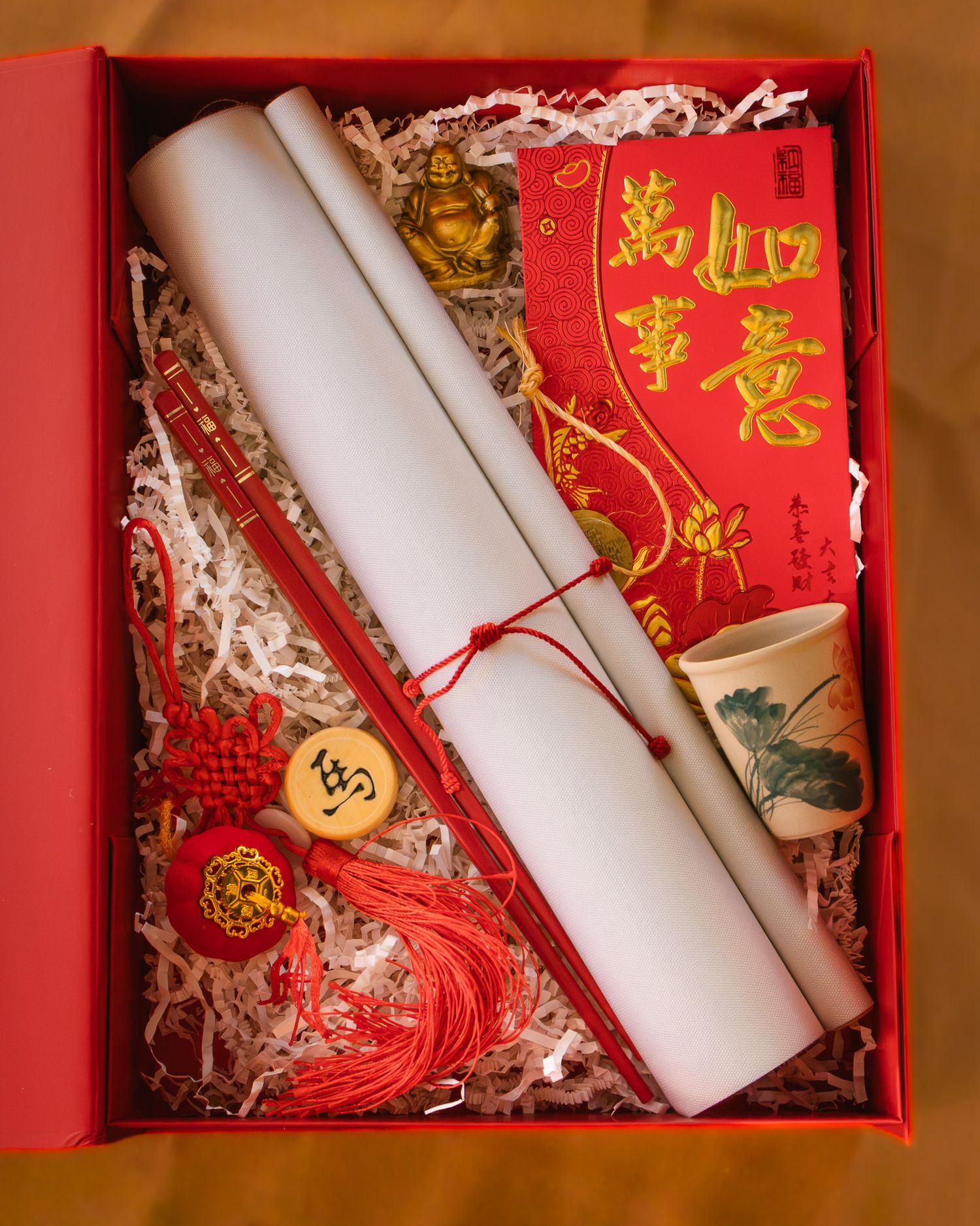 Red gift box with a rolled scroll, red envelope, chopsticks, ceramic teacup, Chinese knot tassel charms, and a golden Buddha figurine.