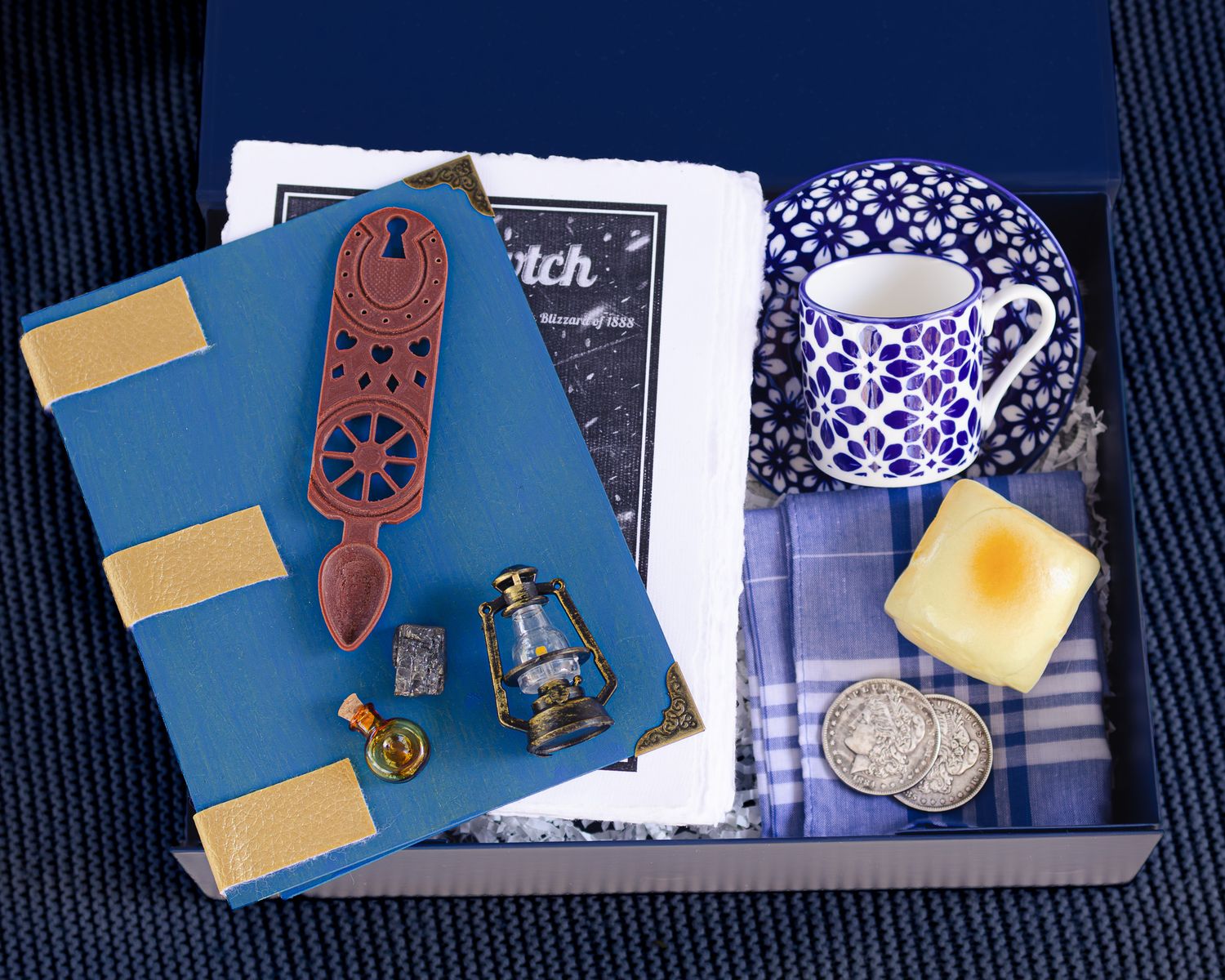 Gift box with blue handmade journal, carved wooden love spoon, mini lantern, blue patterned teacup, plaid cloth, soap bar and silver coins.