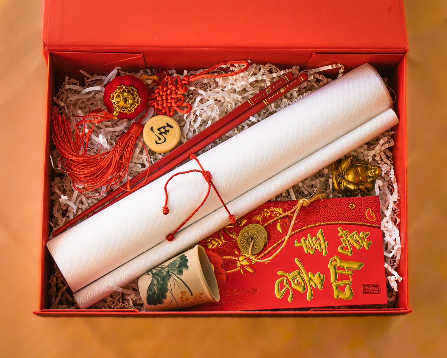 Open red gift box with a rolled white scroll, red Chinese knot charm, lucky red envelope and gold coin accents on white paper shred filler.