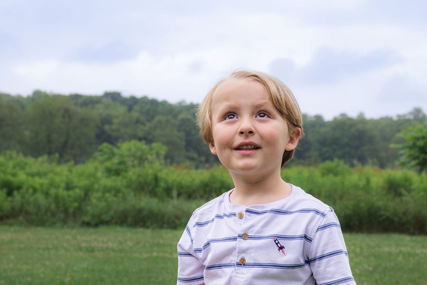 Adorable blonde toddler boy smiling and looking up in a green outdoor park setting.