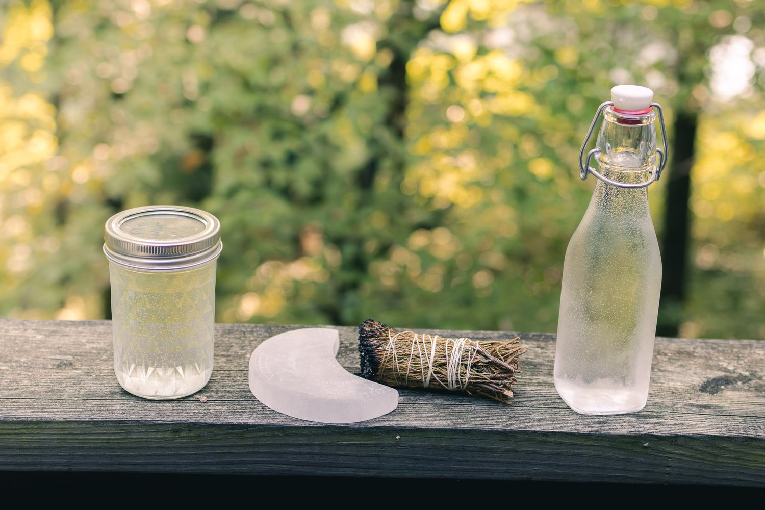 Mason jar, glass bottle, and sage bundle arranged on a rustic wooden railing outdoors.