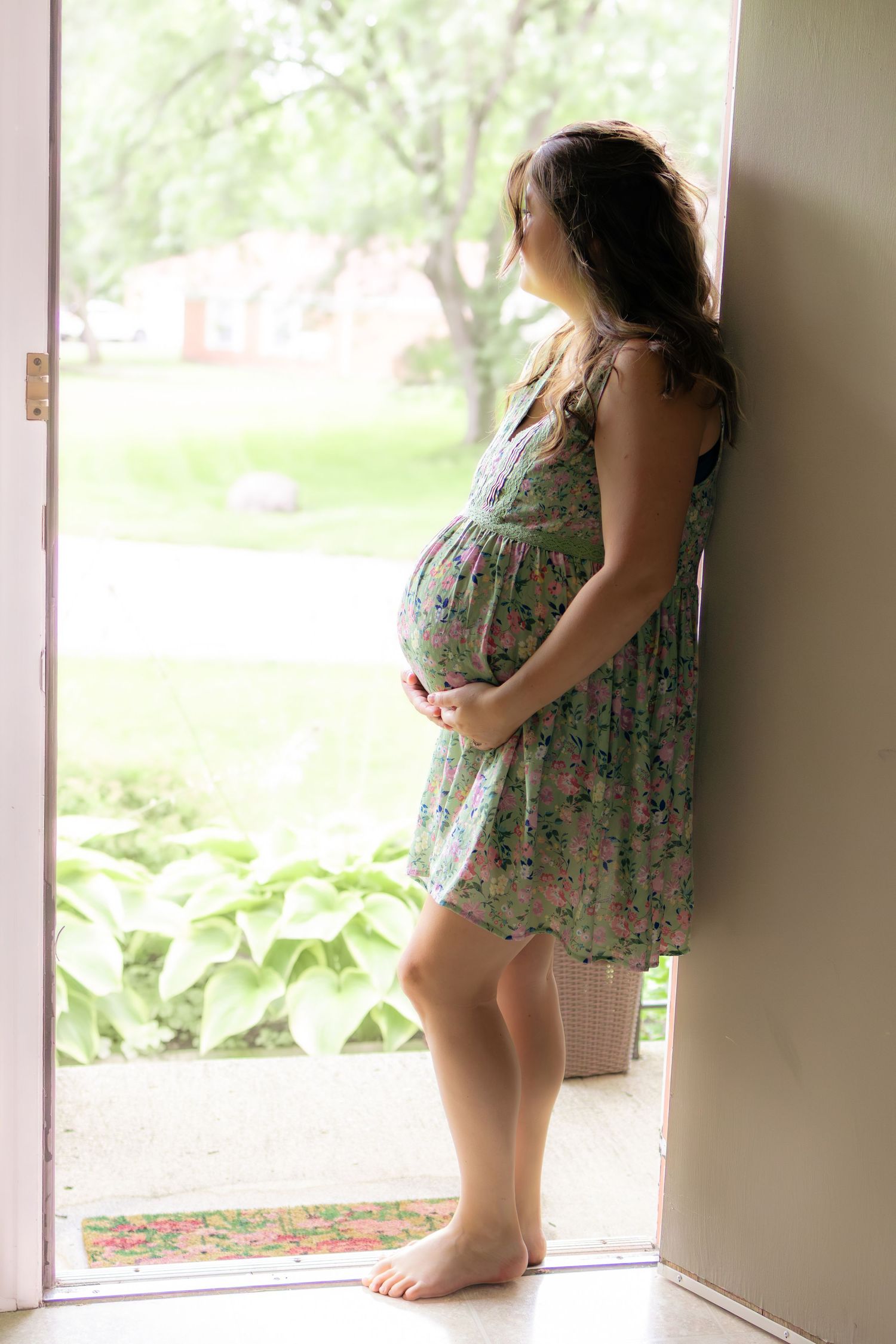 Pregnant woman in floral dress standing barefoot in open doorway looking outside.