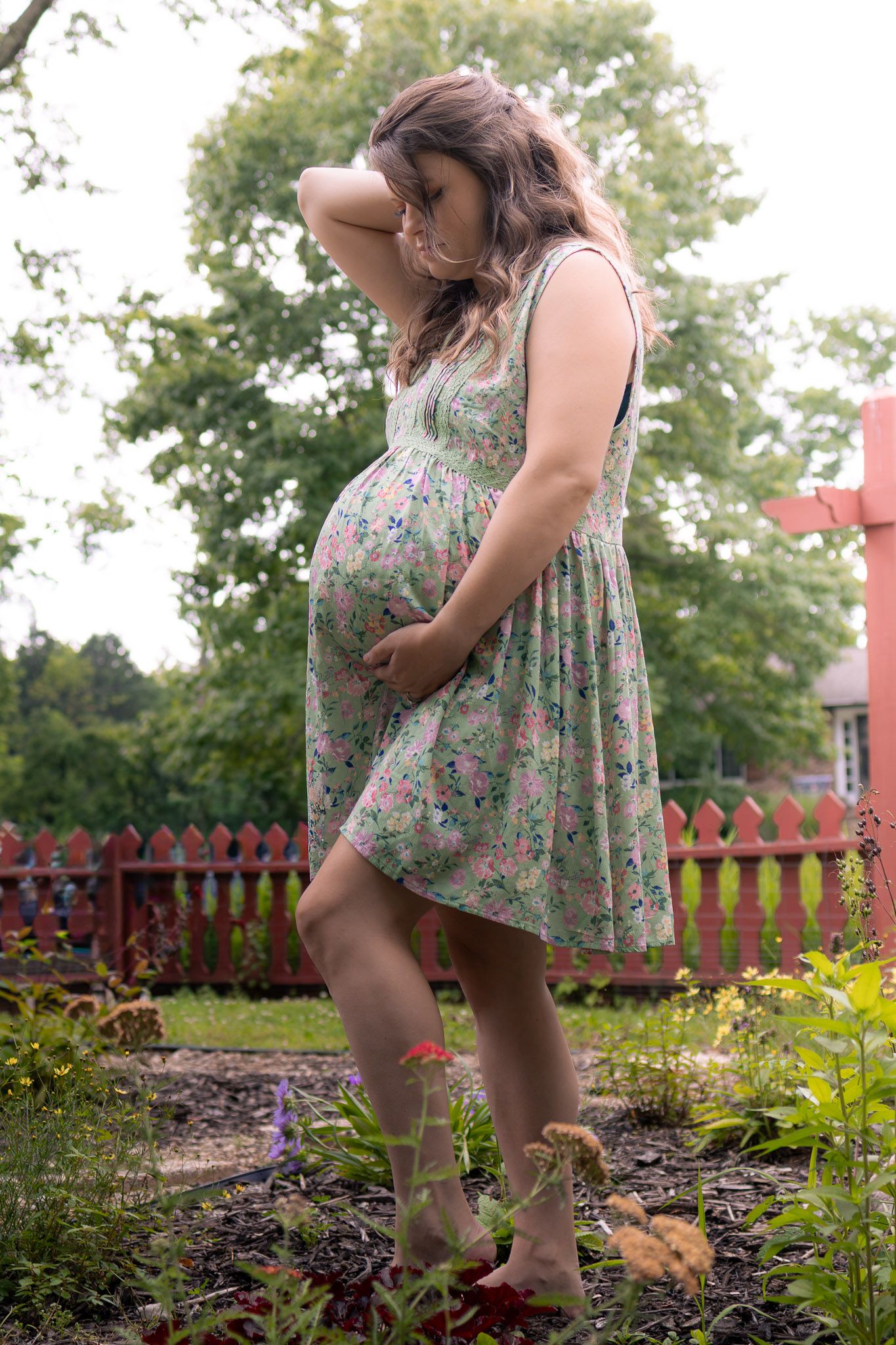 Barefoot pregnant woman in floral dress standing in garden holding her baby bump.