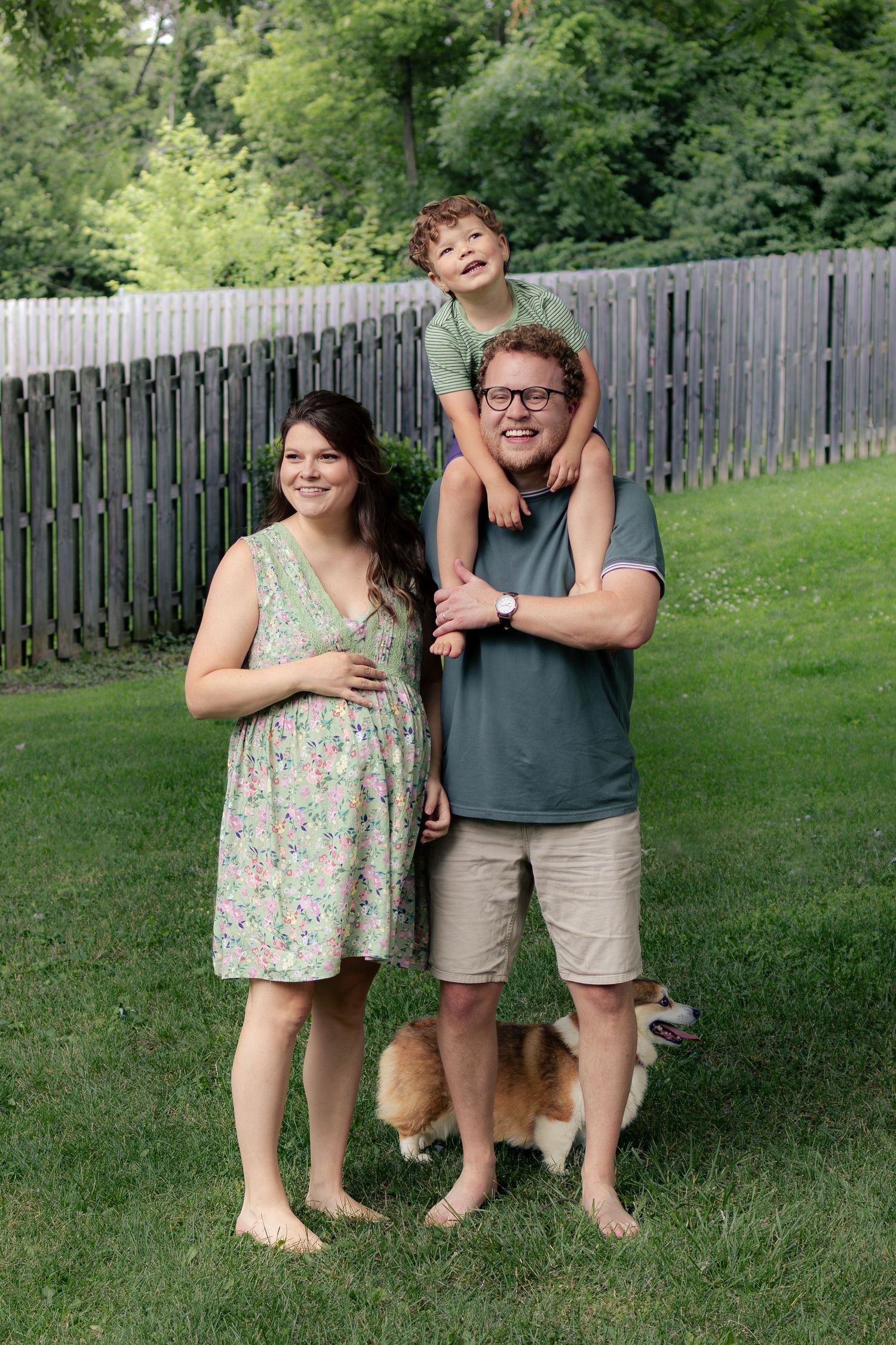 Happy pregnant family with young boy on dad's shoulders and corgi dog posing in backyard.
