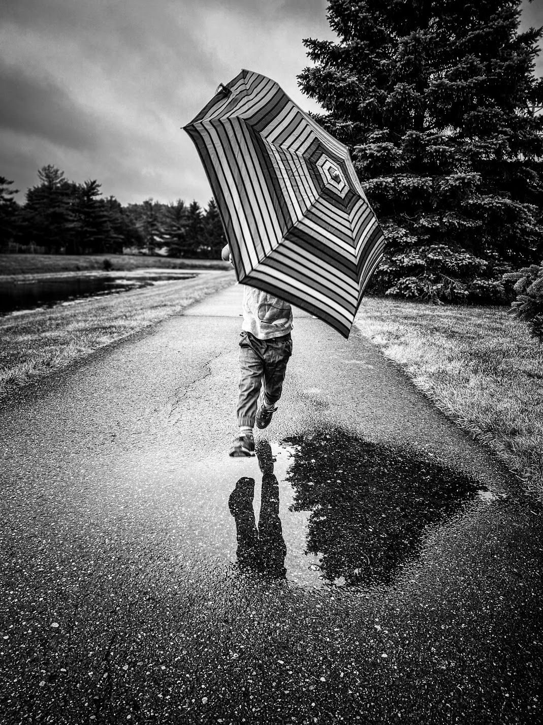 A child runs through a rainy street holding a striped umbrella, reflected in a puddle below in black and white.