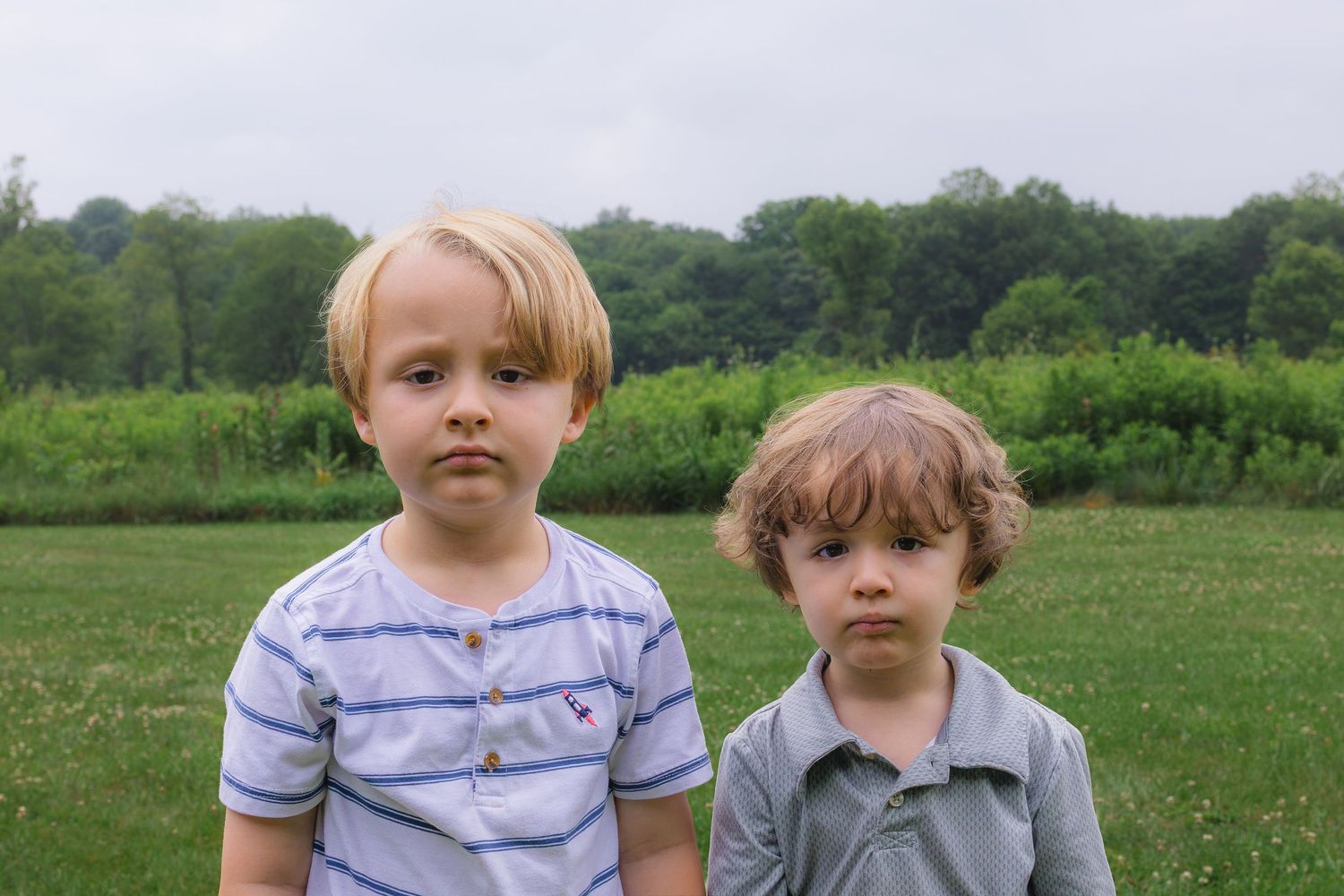 Two young blonde boys standing together in a green field with trees in the background.
