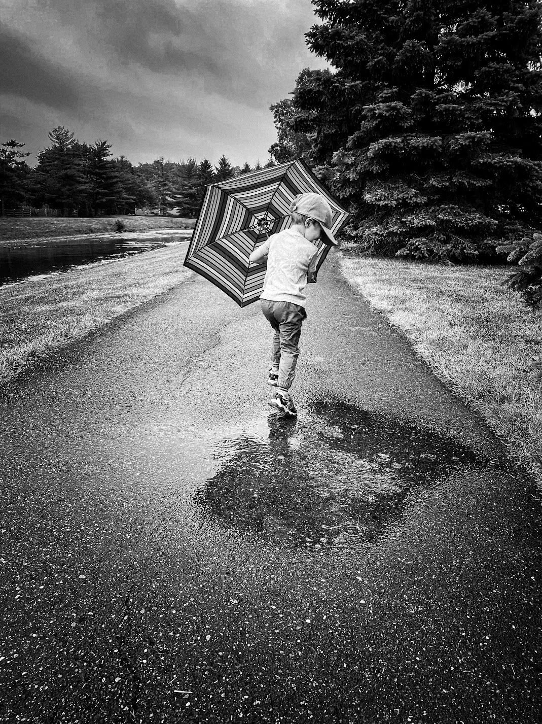 A child jumping in rain puddles while holding a striped umbrella on a cloudy day.
