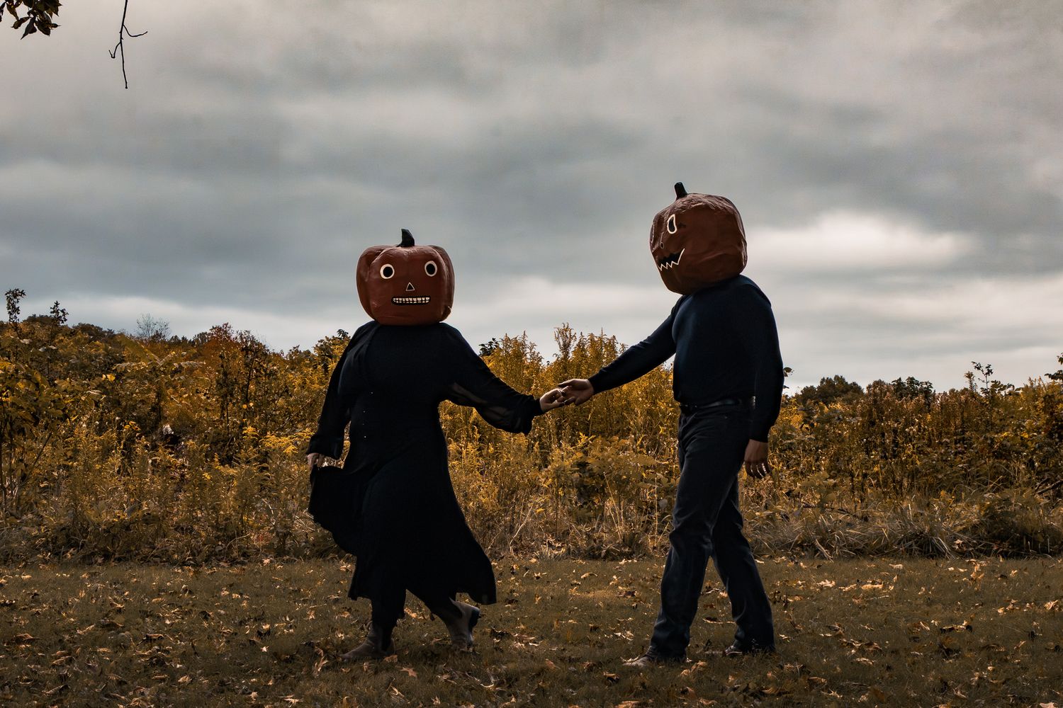 Two people wearing jack-o-lantern pumpkin heads holding hands in a field on a cloudy autumn day.