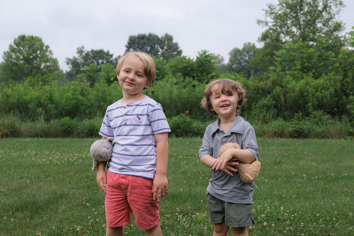 Two young boys smiling and holding stuffed animals while standing in a green meadow.