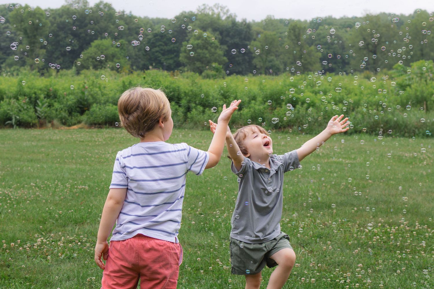 Two young boys reaching up to catch soap bubbles together in a green outdoor field.