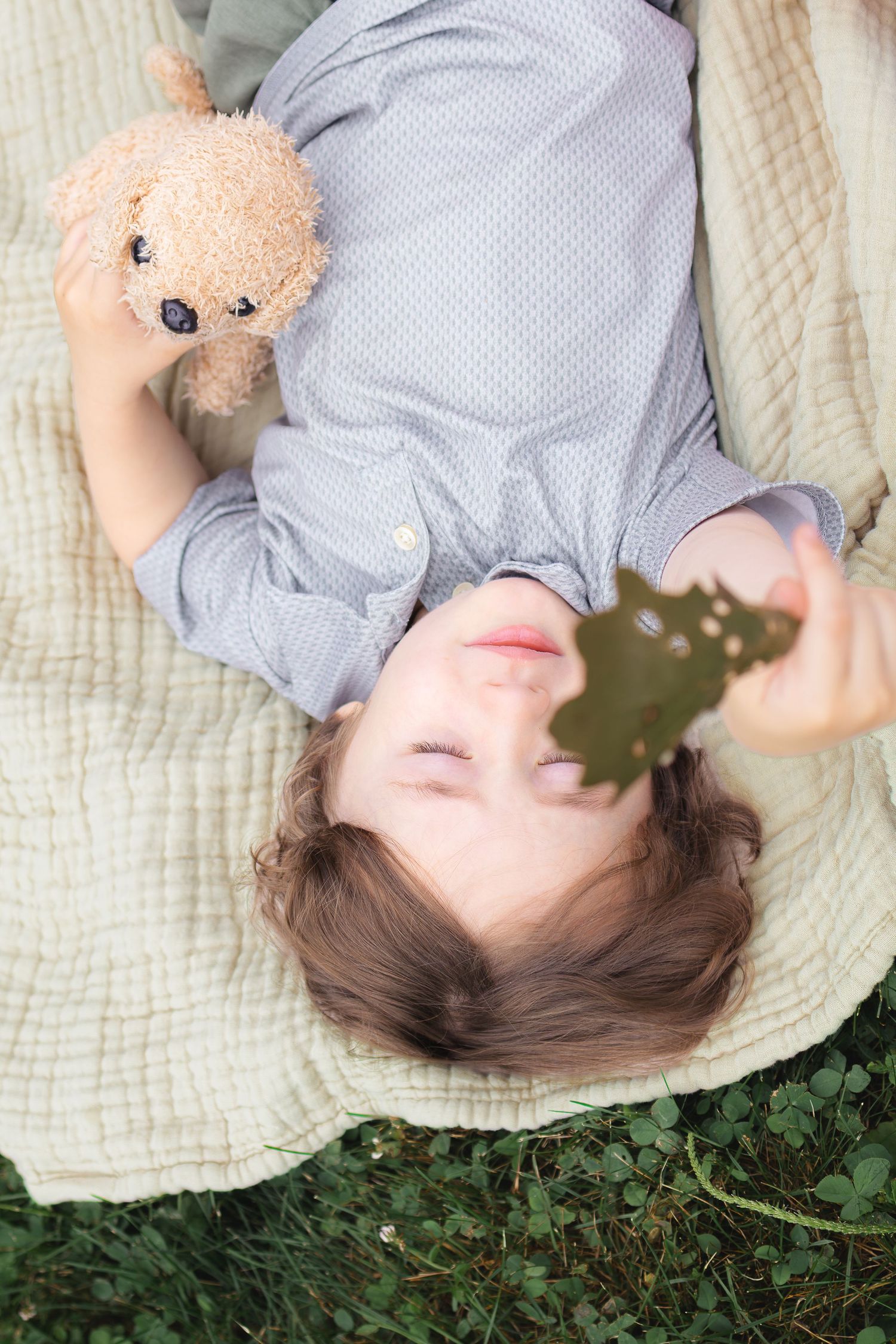 A young boy lies on a blanket in the grass, smiling while holding a teddy bear and a leaf.